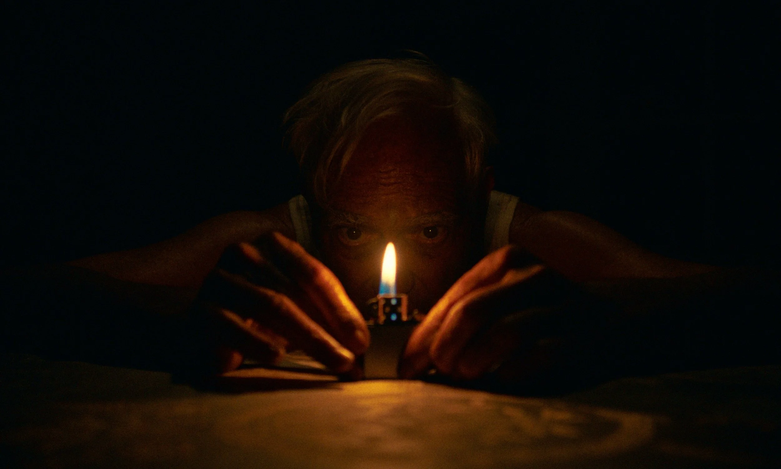 Person with light-colored hair and wrinkled skin looks directly at camera in low light, holding a lit matchstick close to the camera.