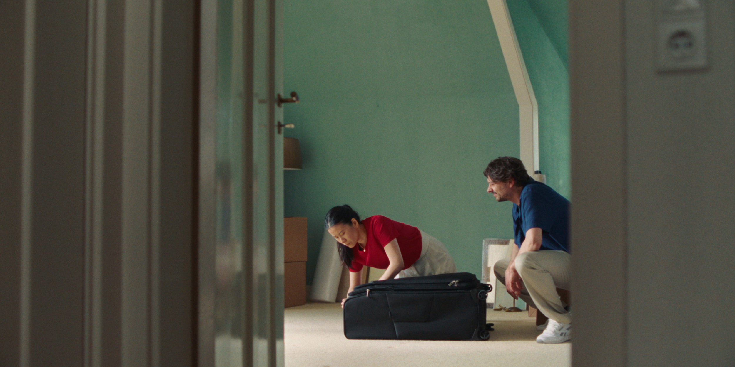 A man and woman are packing a black suitcase in a green-walled room, seen from a doorway.