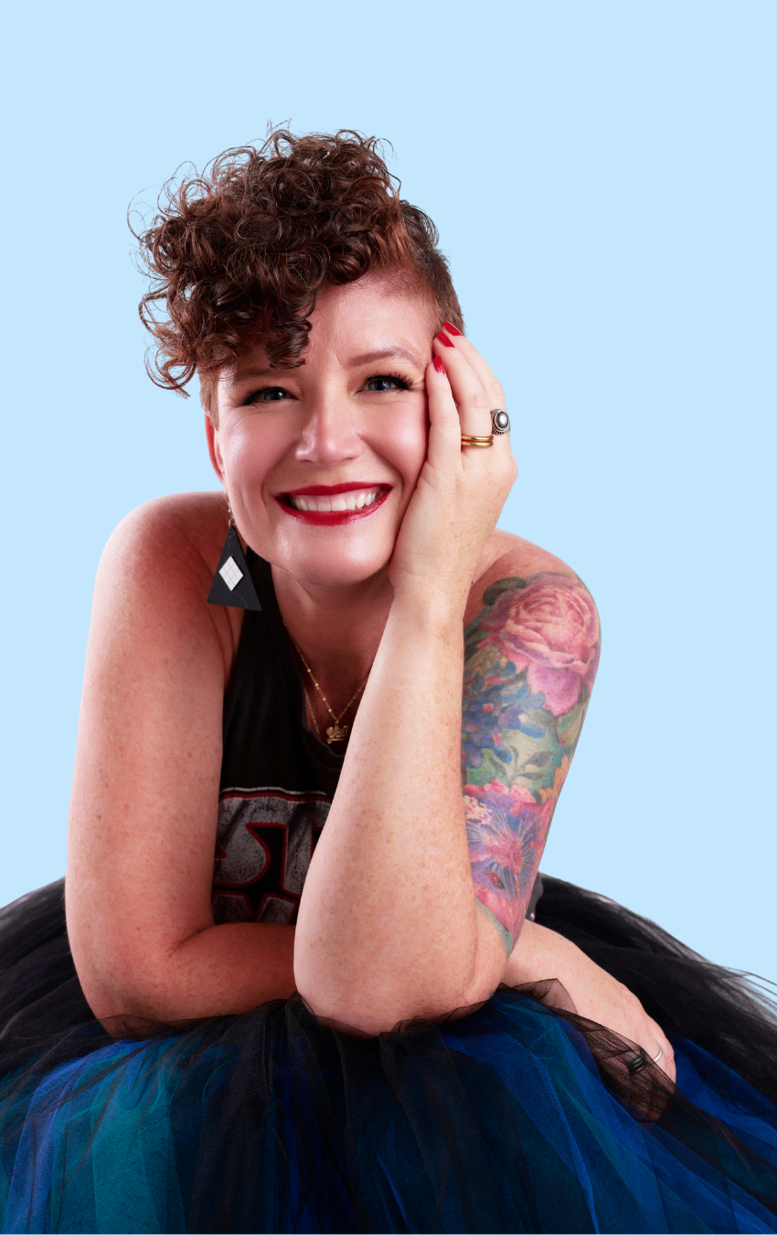 A smiling woman with short curly hair, tattoos on her arm, wearing a black tank top and a colorful tulle skirt, posed against a light blue background.