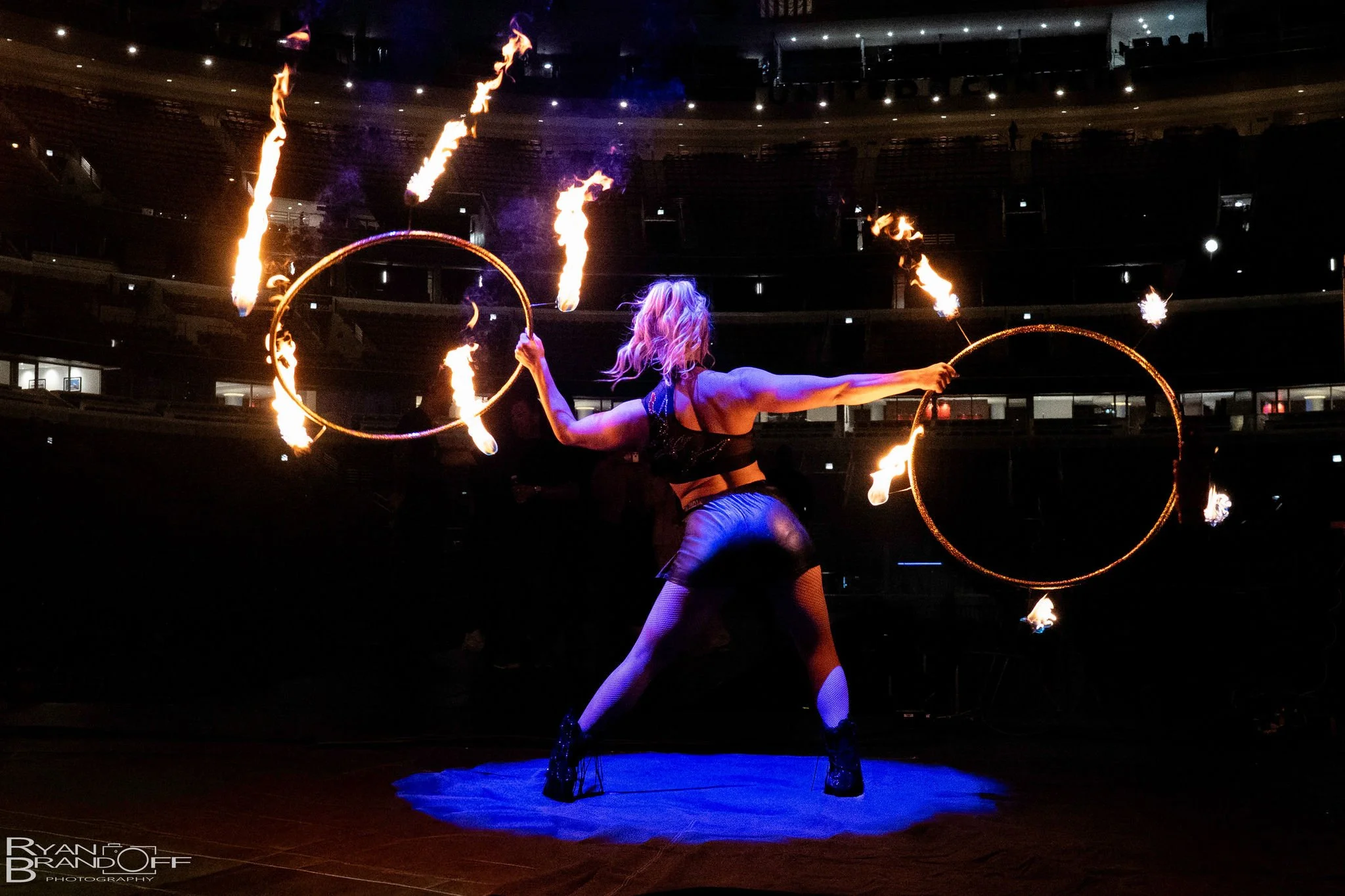 A female fire performer spinning flaming hoops on stage in a dark theater, with an audience in the background.