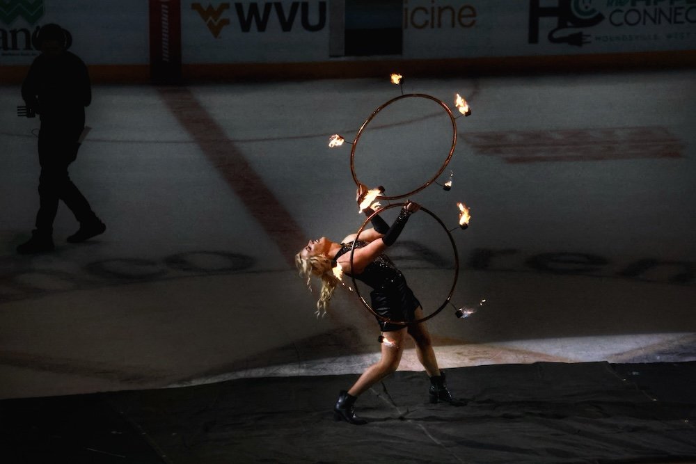 A female fire performer spinning two flaming hula hoops at an ice hockey arena.