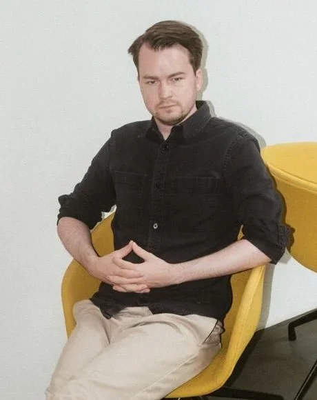 A young man sits on a yellow chair against a white background, wearing a black jacket and beige trousers.