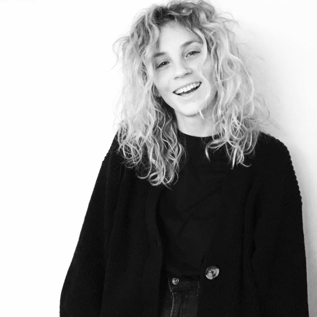 A smiling young woman with curly hair, wearing a black sweater, photographed against a white background.