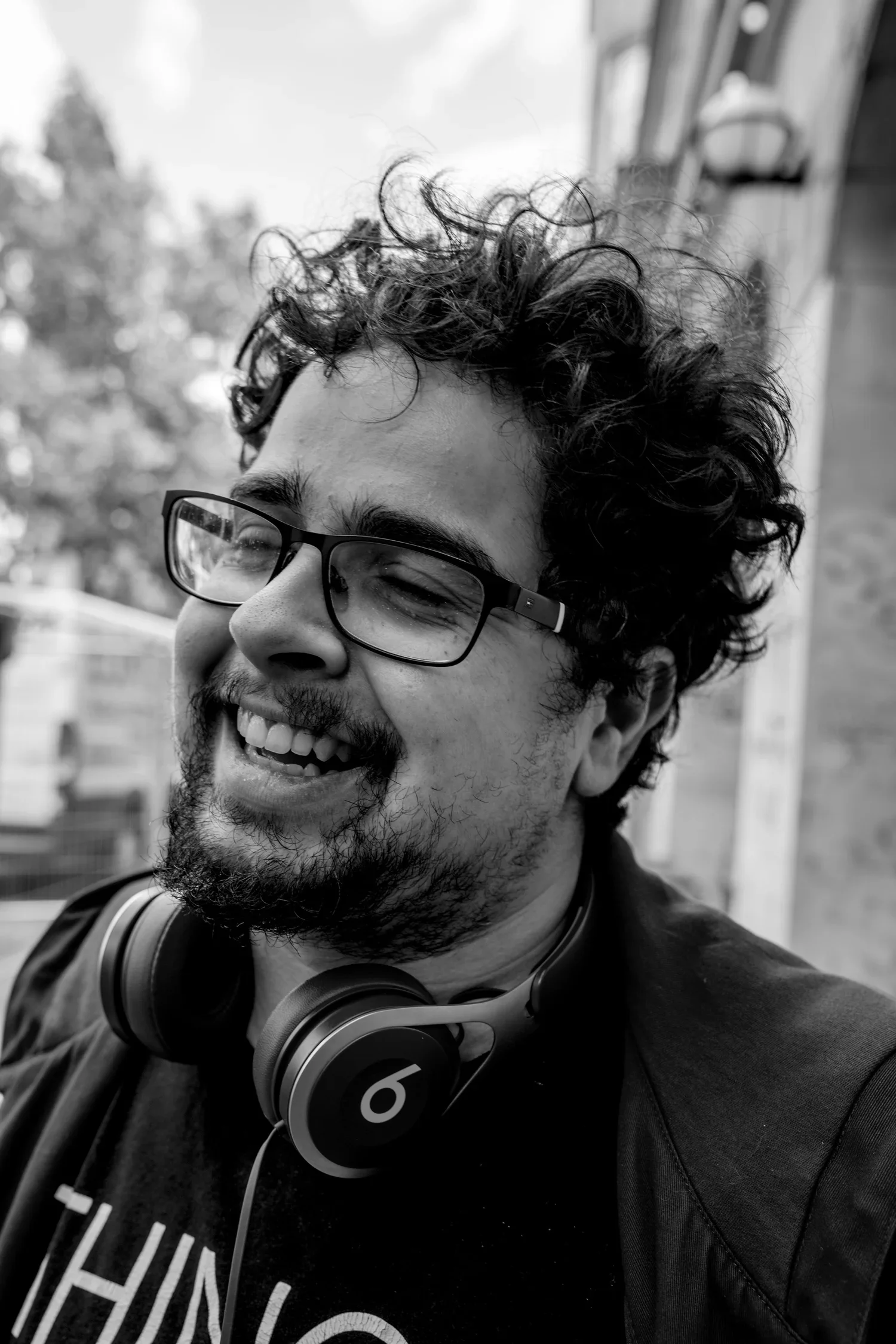 A laughing man with glasses, curly hair and a beard, wearing headphones around his neck, outdoors on a cloudy day.