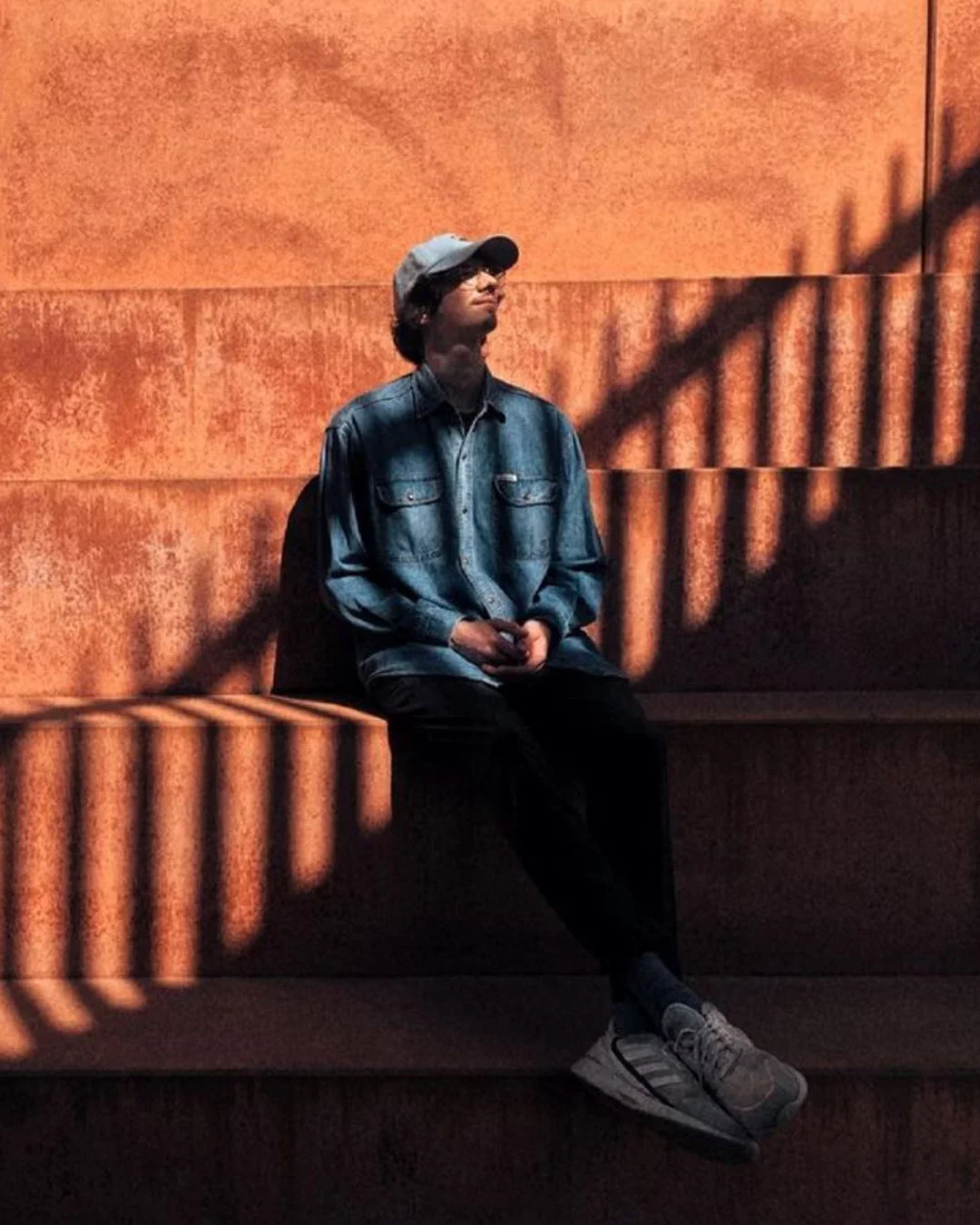 A young man sits on a bench in front of a wall of mixed orange-brown tones and shadows. He is wearing a cap, a blue denim jacket, black trousers and sneakers.