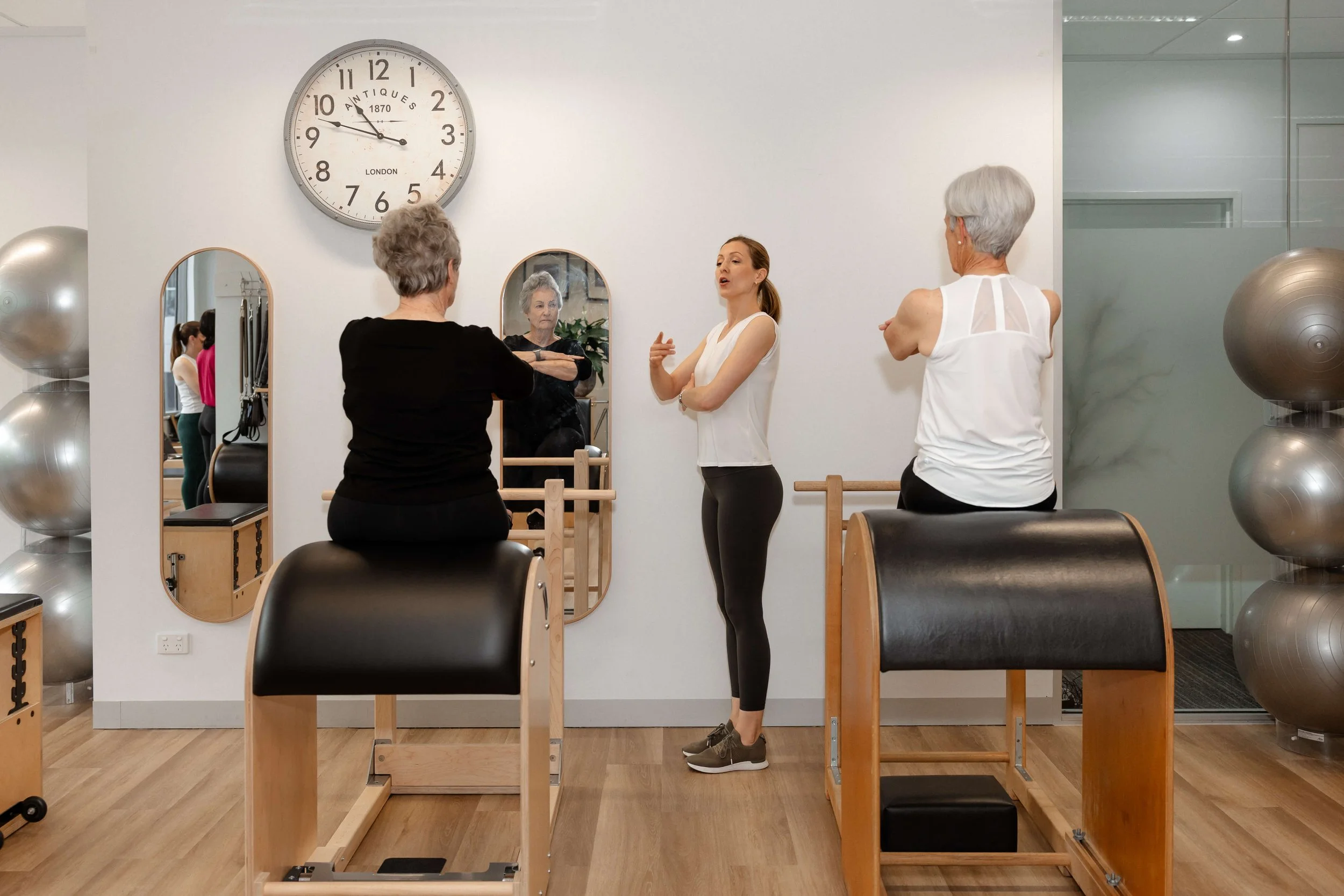 A fitness instructor instructs two older women during a Pilates class in a gym studio with mirrors, exercise equipment, and large silver exercise balls.