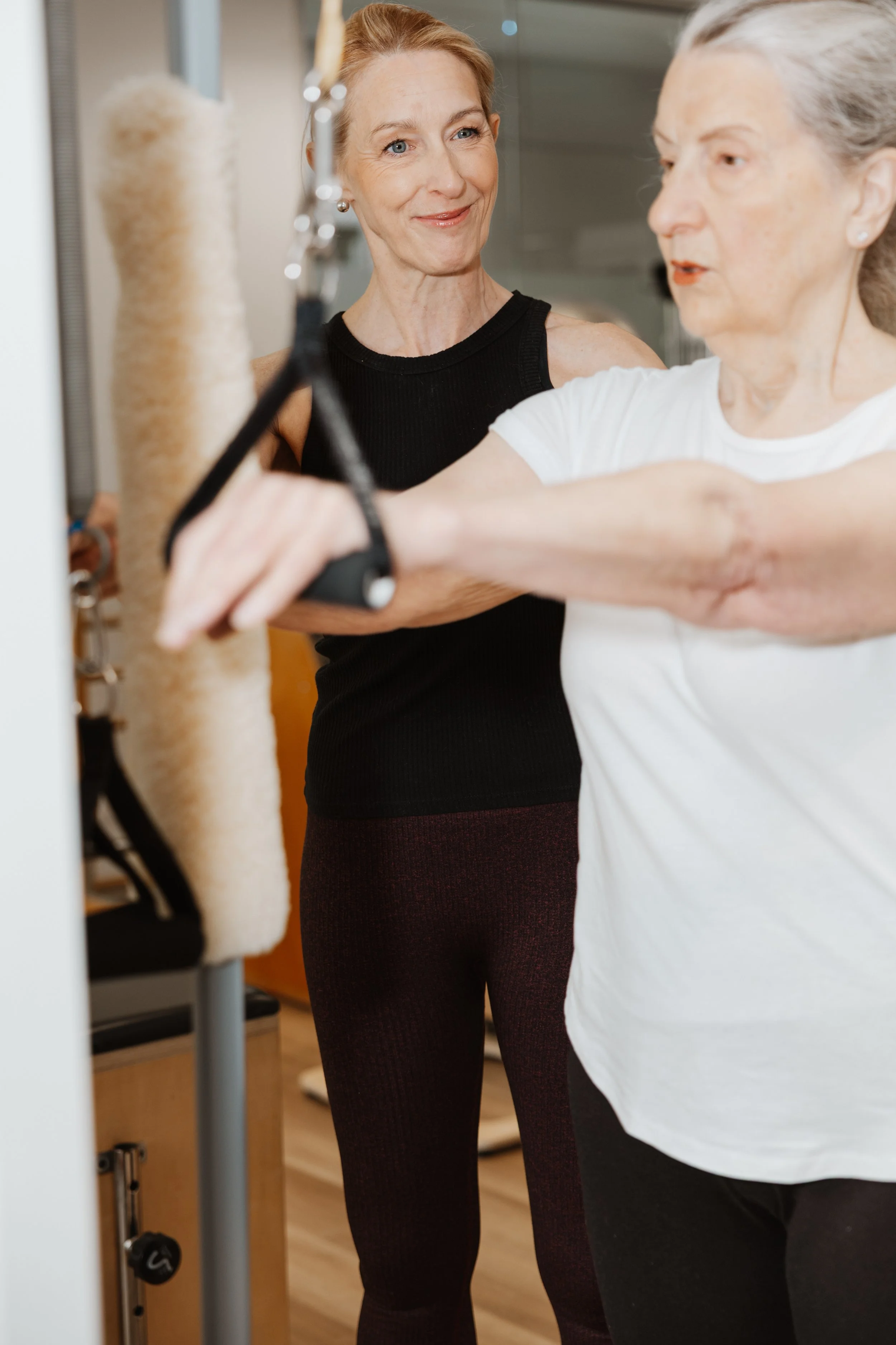 Two women, one younger and one older, are exercising with a resistance band in a gym or physical therapy setting. The older woman is holding the band and the younger woman appears to be instructing or assisting her.