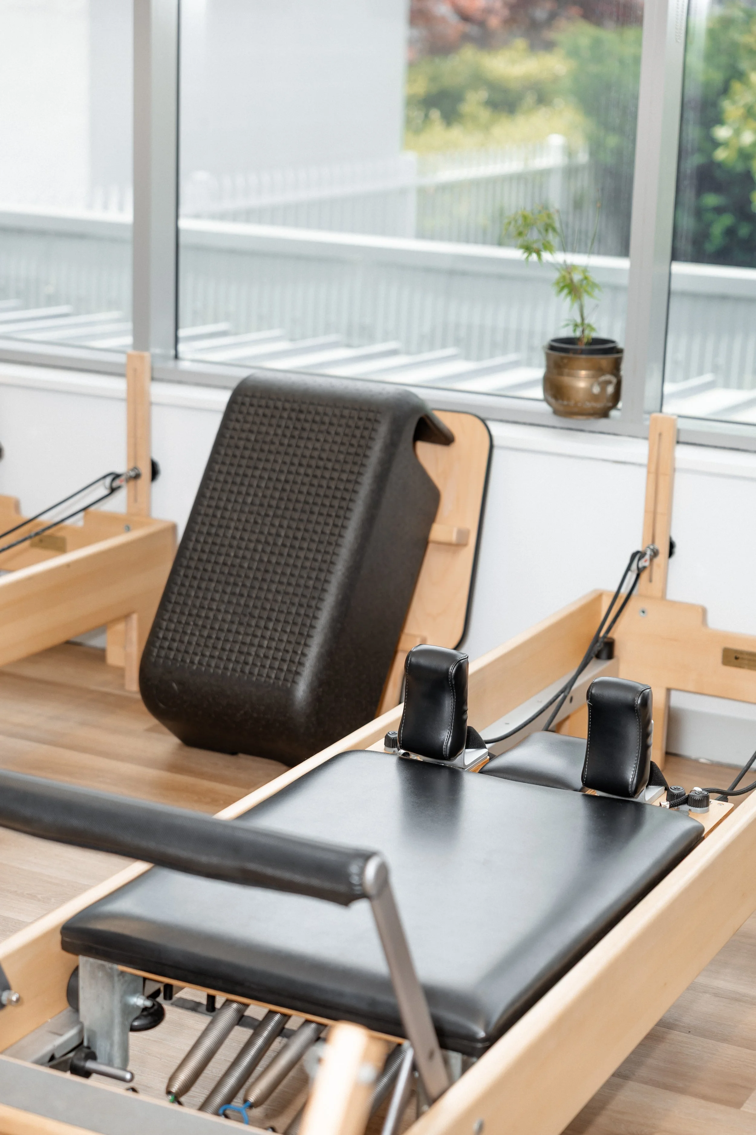 A Pilates reformer machine with a black cushioned platform and headrests in a fitness studio, near a window with a small potted plant on the sill.