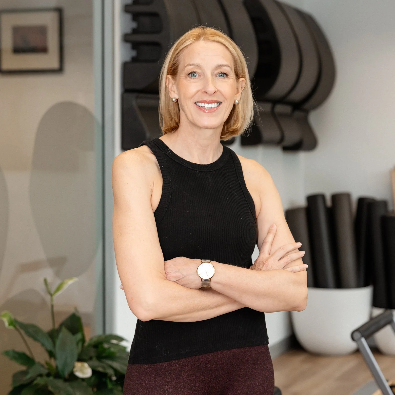 A woman with blonde hair, wearing a black sleeveless top and a watch, smiling with her arms crossed in a fitness studio or gym setting.