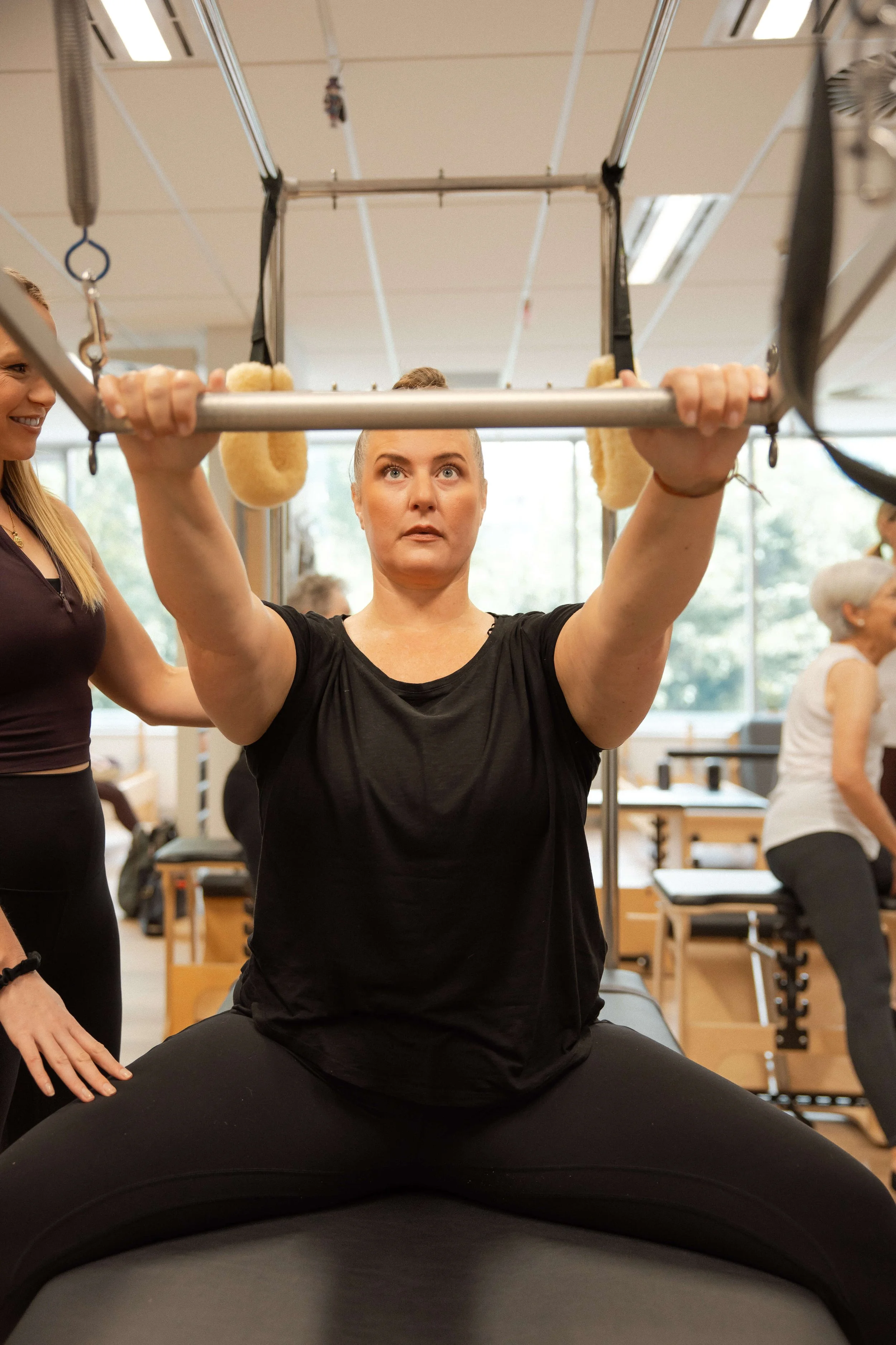 A woman performing a squat on an exercise machine, with one woman assisting and others in the background in a gym or physical therapy setting.