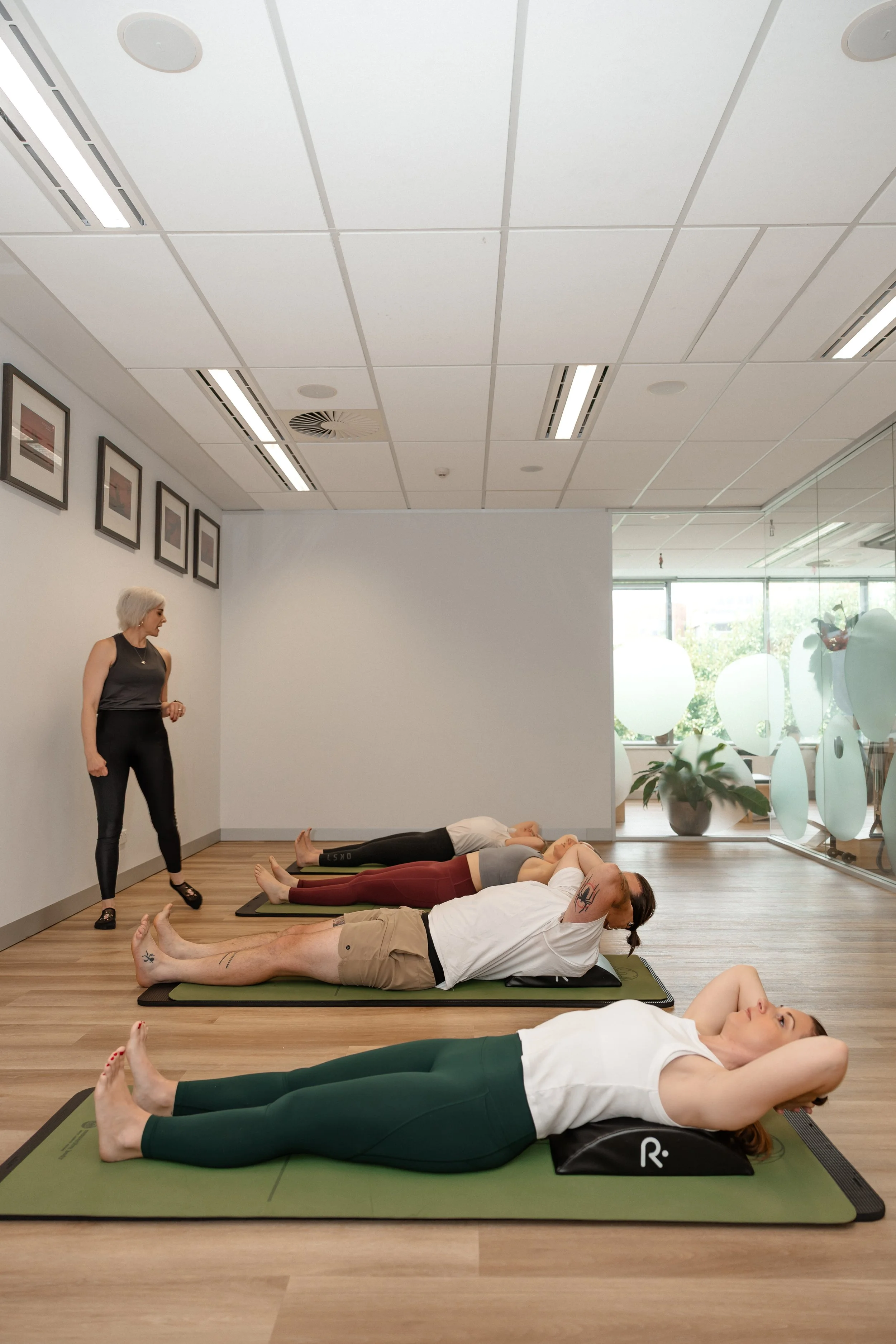 A group yoga class with four people lying on yoga mats in a modern studio, with a woman instructor standing nearby.