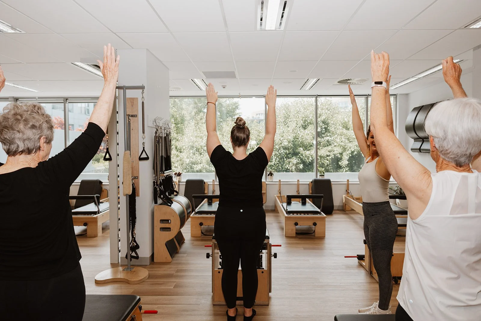 Group of elderly women and a young instructor participating in a fitness class in a well-lit studio, raising their arms during exercise.
