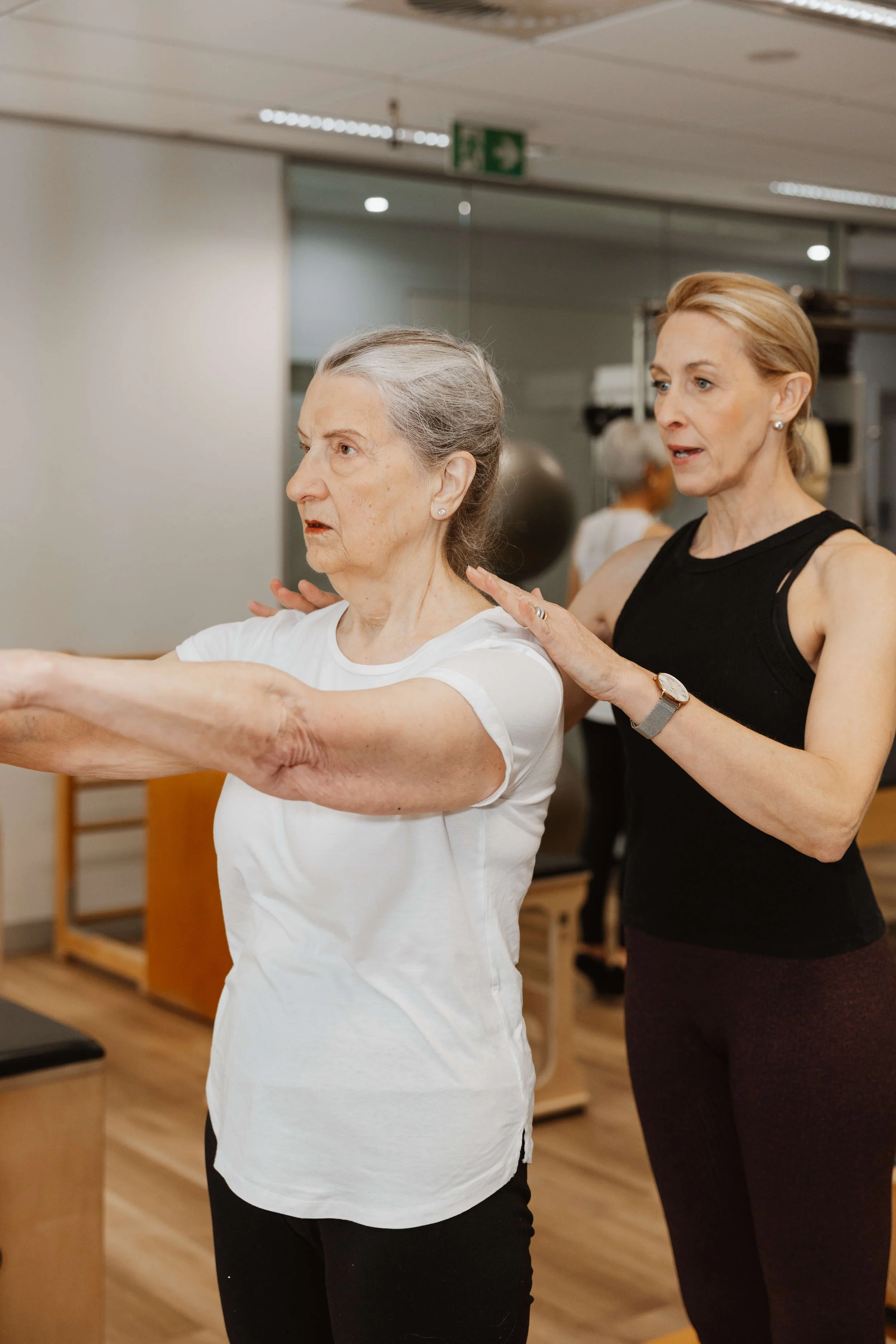 An elderly woman with gray hair doing physical therapy exercises while a female trainer assists her in a gym or rehabilitation center.