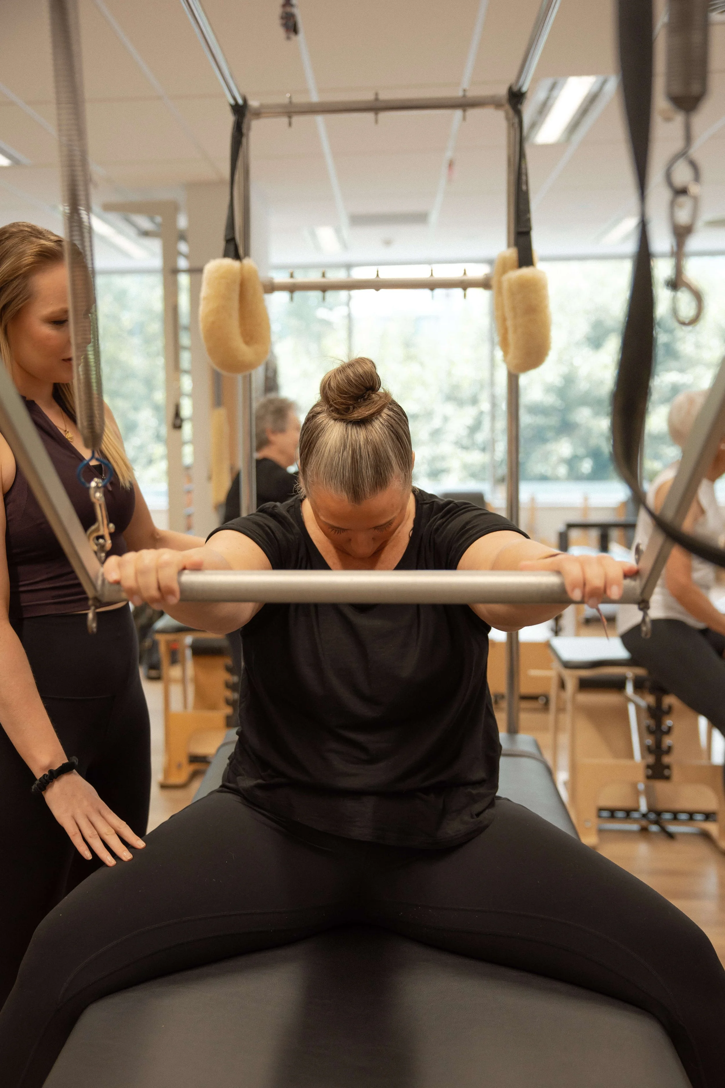 A woman practicing a squat exercise with a barbell at a physical therapy or rehabilitation center, guided by a therapist, with other people in the background.