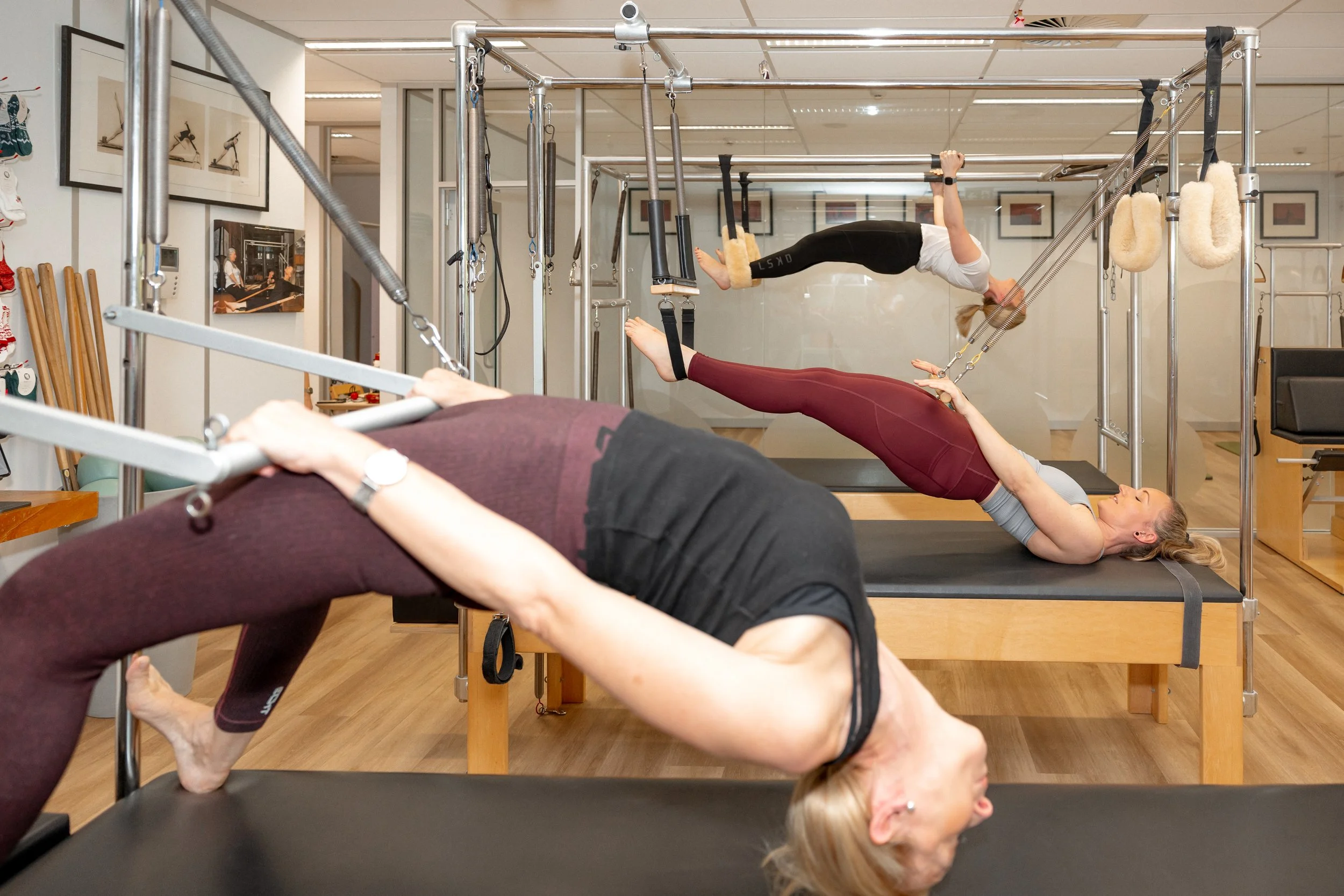 Two women exercising on Pilates reformer machines in a Pilates studio. The woman in the foreground is lying on her back, performing a bridge exercise, while the woman in the background is hanging upside down, practicing suspension exercises.