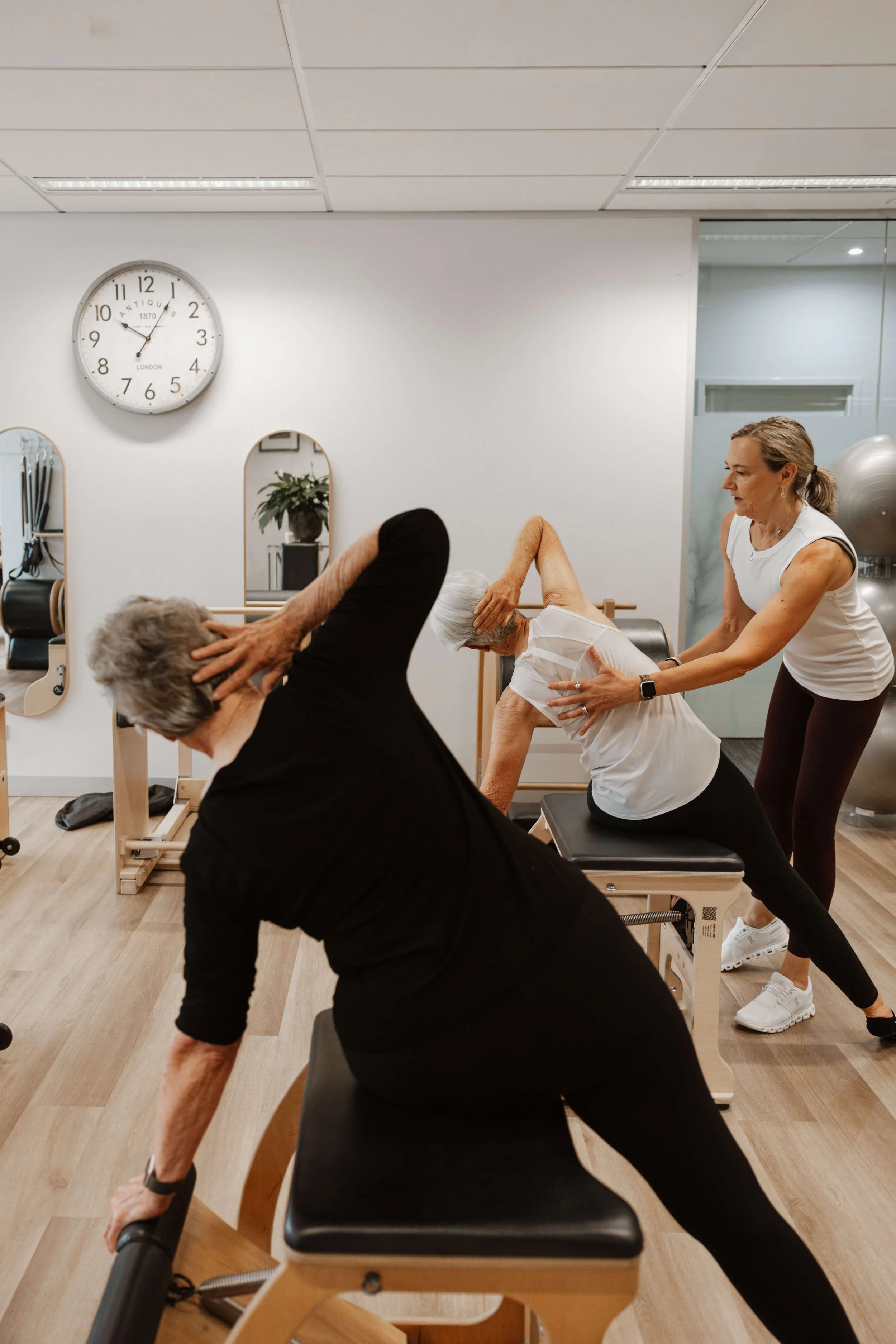 A beige-haired elderly woman stretches with assistance from a trainer during a physical therapy session in a gym.