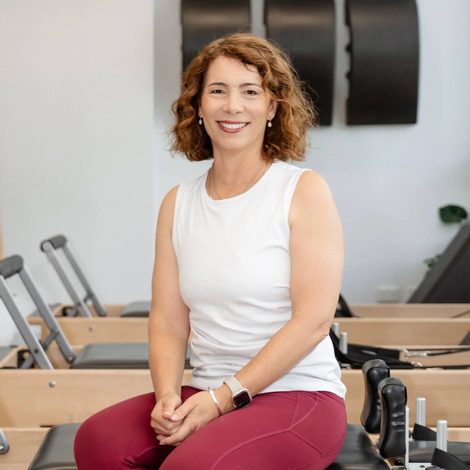 A woman with curly red hair smiling, sitting on a Pilates reformer machine in a fitness studio.