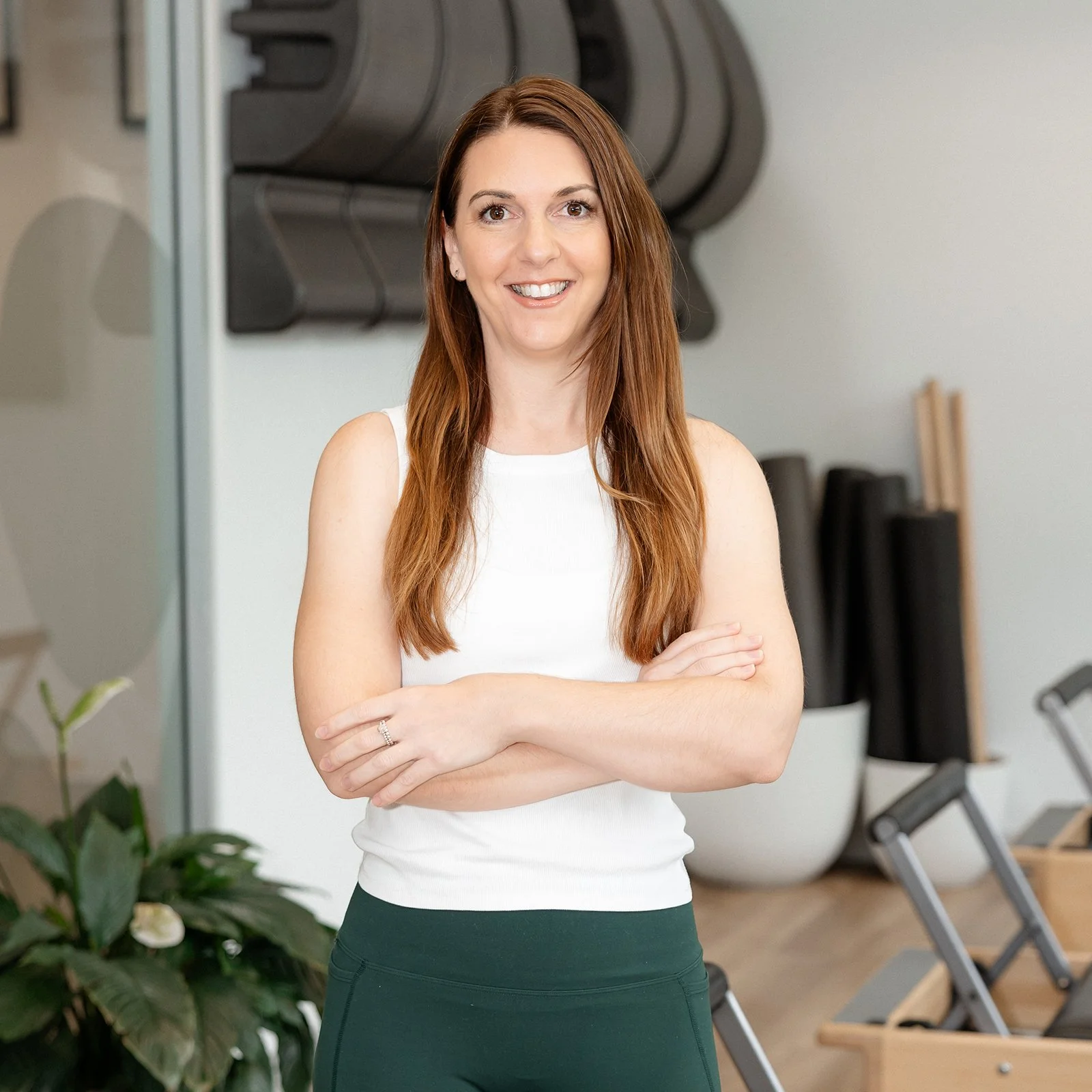 A woman with long red hair smiling and crossing her arms in a fitness or yoga studio.