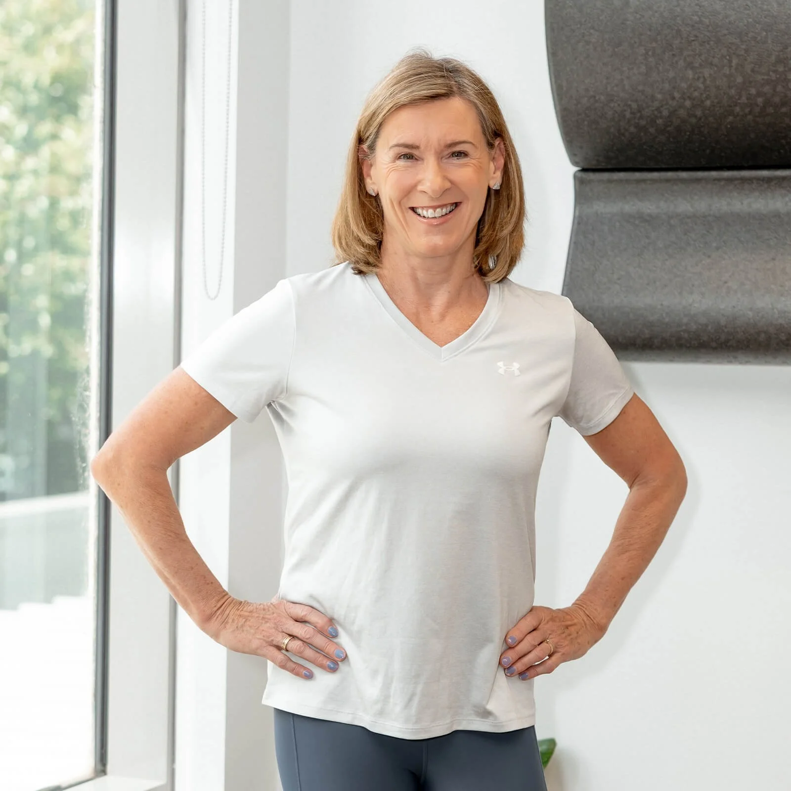 A smiling middle-aged woman with shoulder-length blonde hair, wearing a white athletic t-shirt and gray workout pants, standing indoors with her hands on her hips.