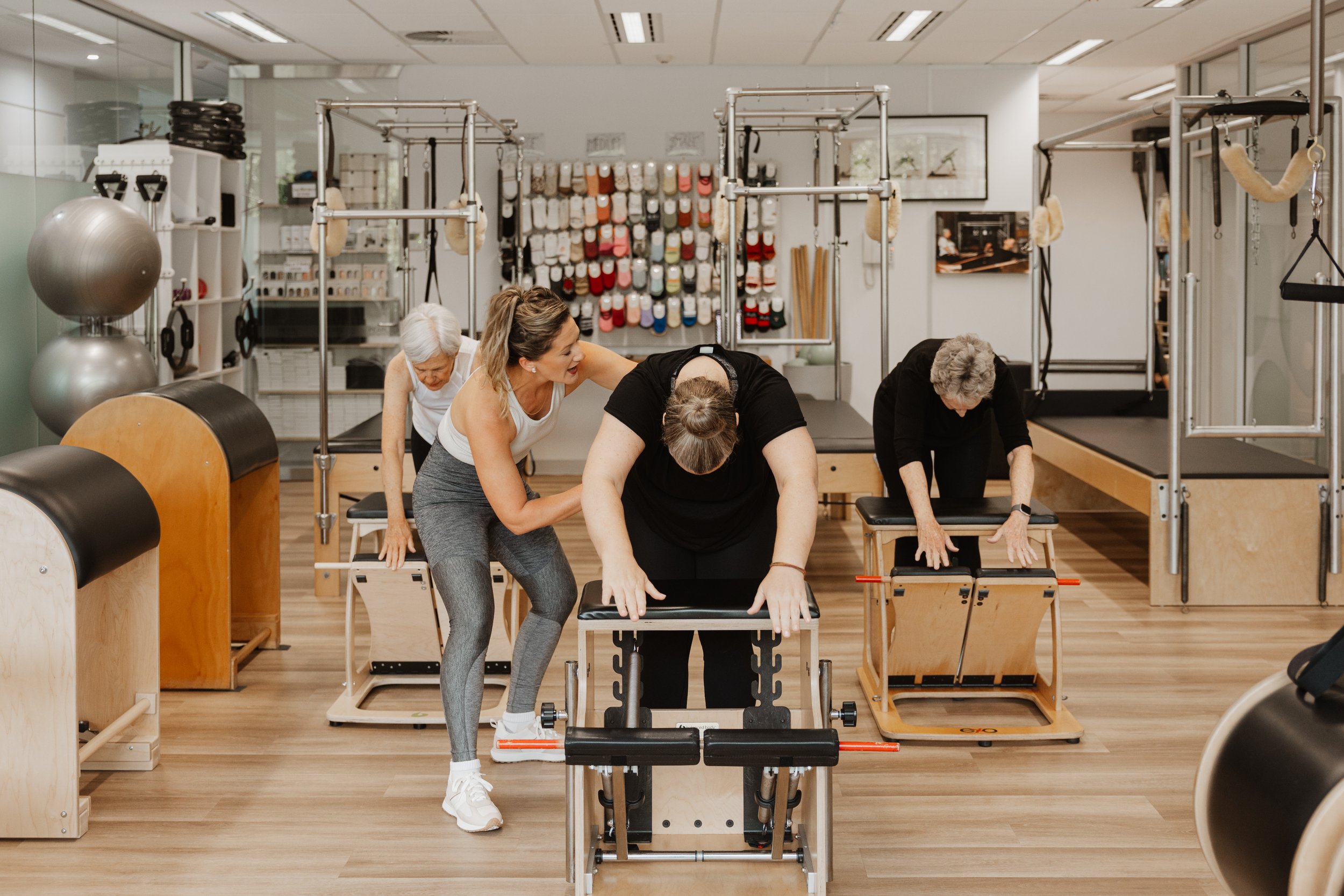 Instructor assisting older women during Pilates class at fitness studio with Pilates equipment, exercise balls, and wall-mounted resistance bands.