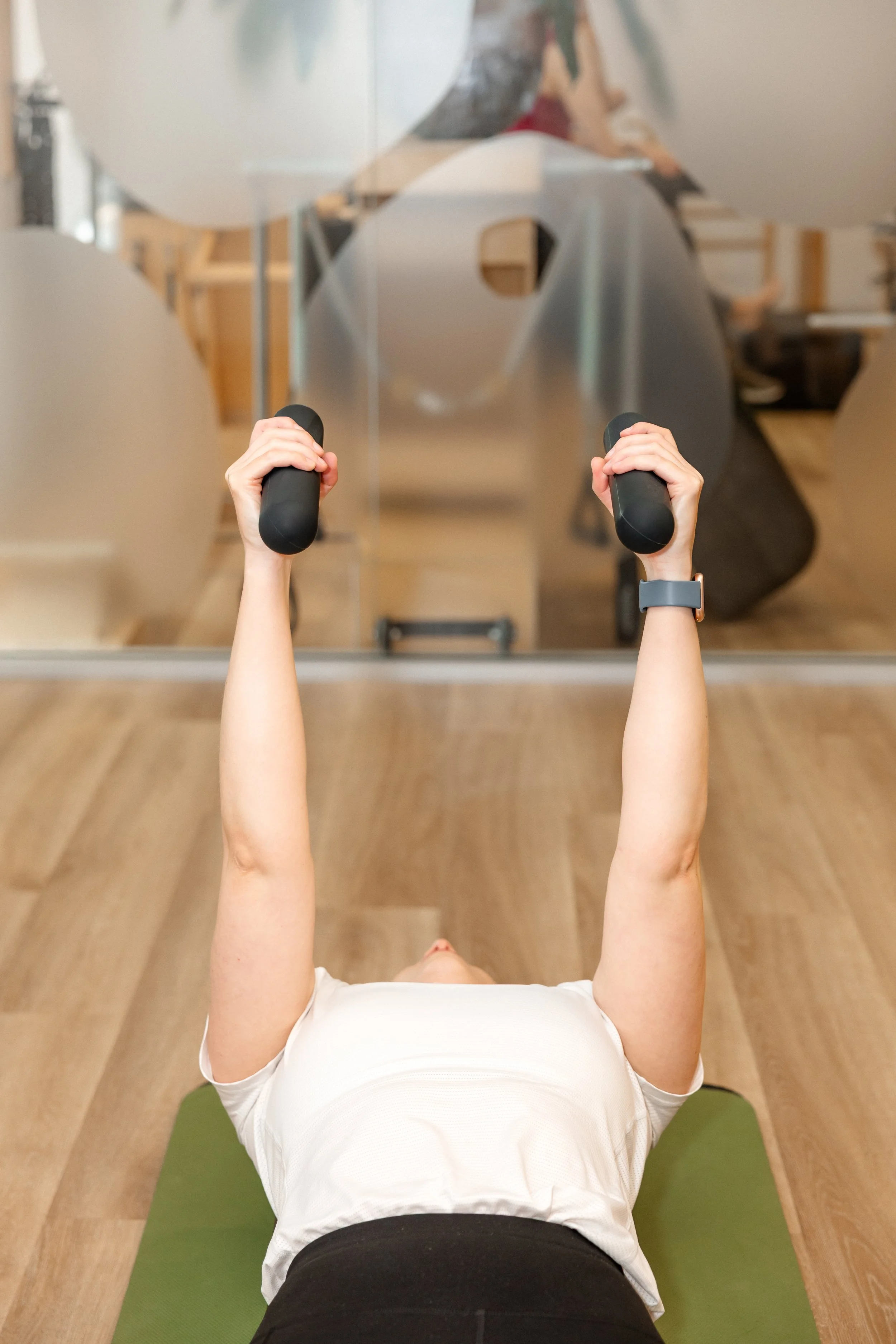 Person lying on a green exercise mat, lifting small dumbbells during a workout in a gym.