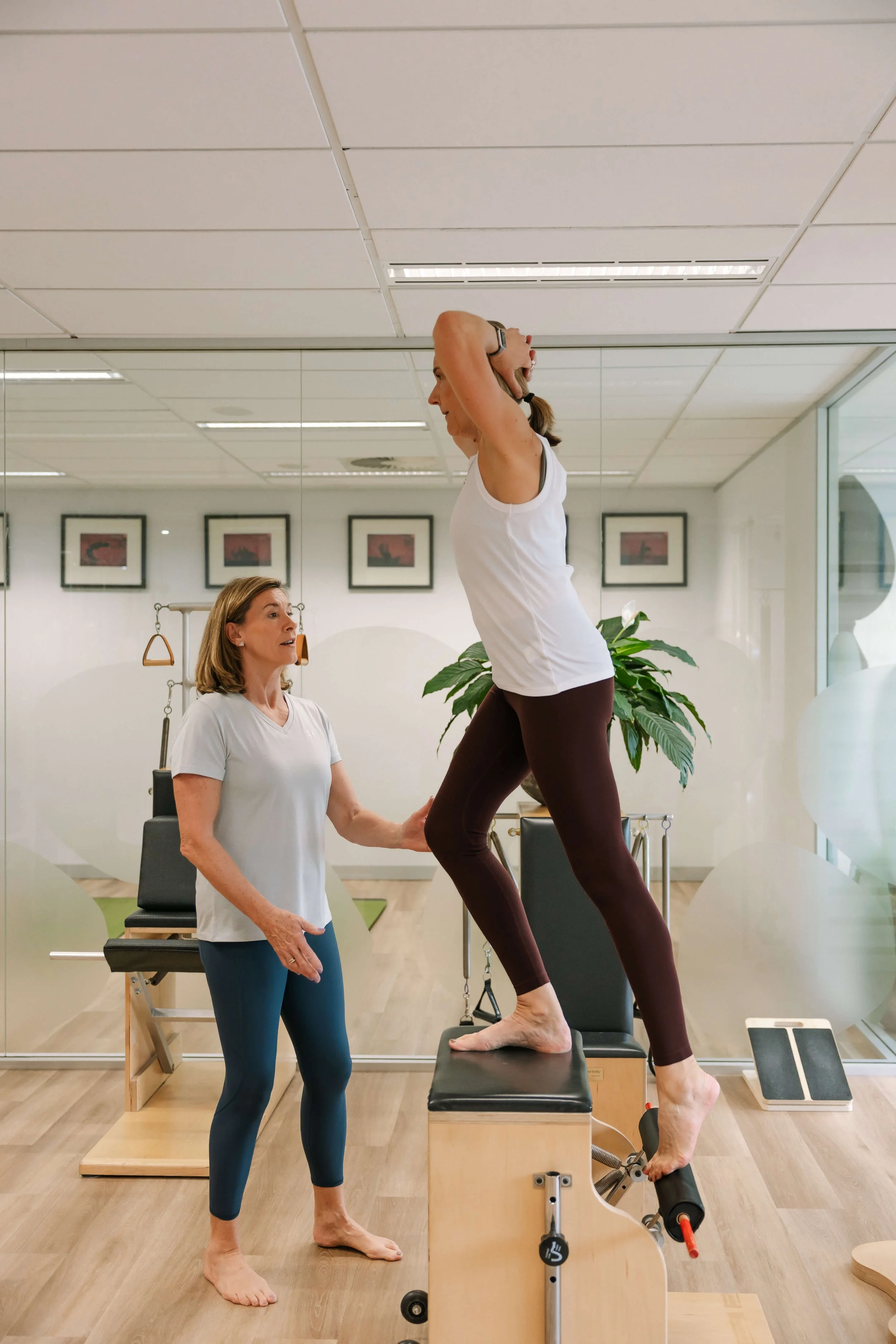 An instructor helping a woman perform a Pilates exercise on a reformer machine in a fitness studio with framed artwork on the wall and a large window.
