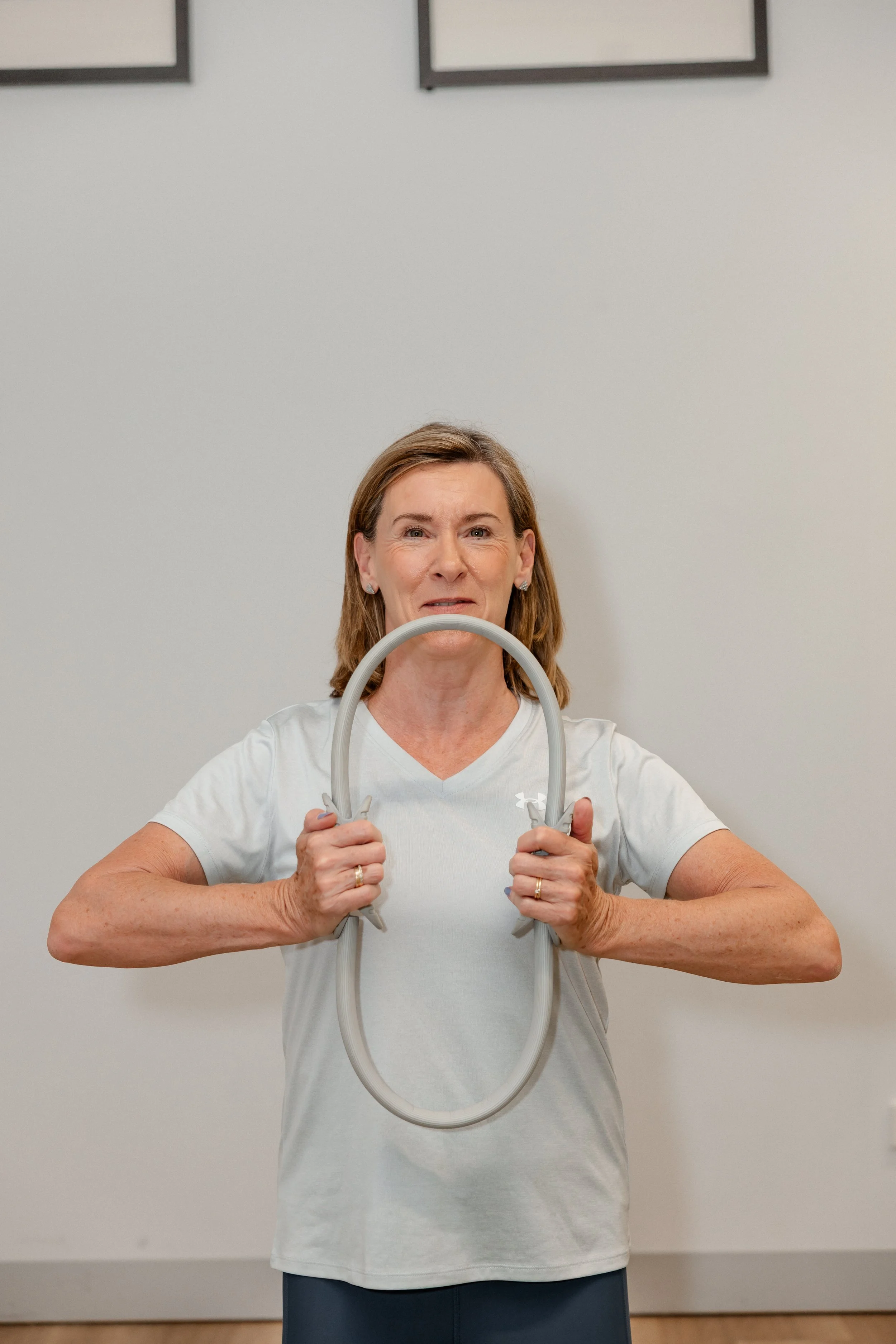 A woman holding a therapy or exercise band in front of her with both hands, standing indoors against a plain wall.