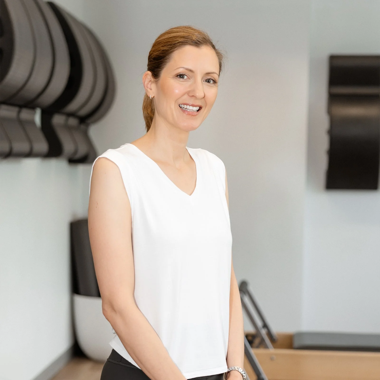 A woman with brown hair in a ponytail, wearing a white sleeveless top, smiling in a modern fitness studio.