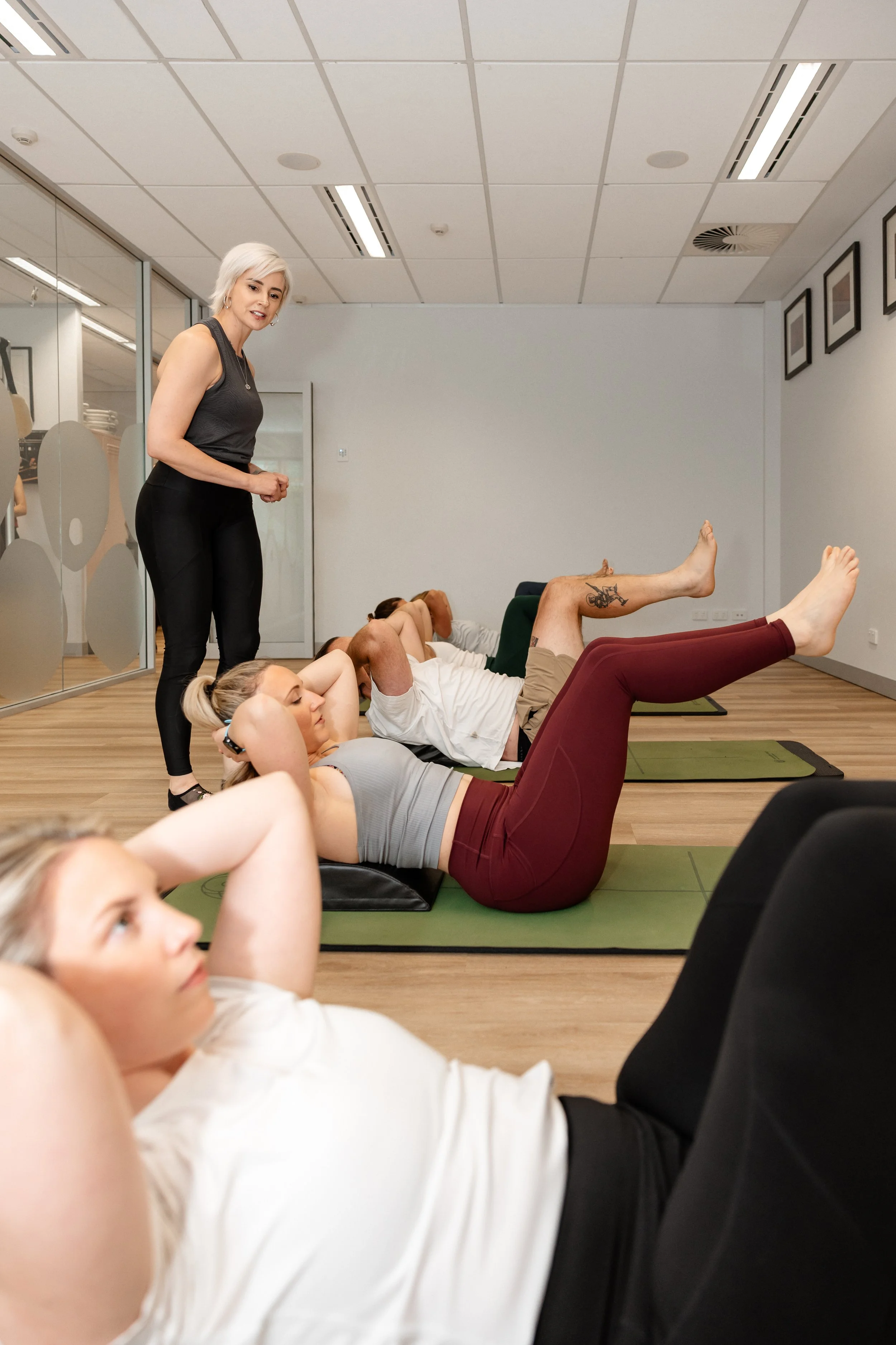 A group fitness class in a studio with women lying on yoga mats and doing abdominal exercises, led by a woman instructor standing nearby.