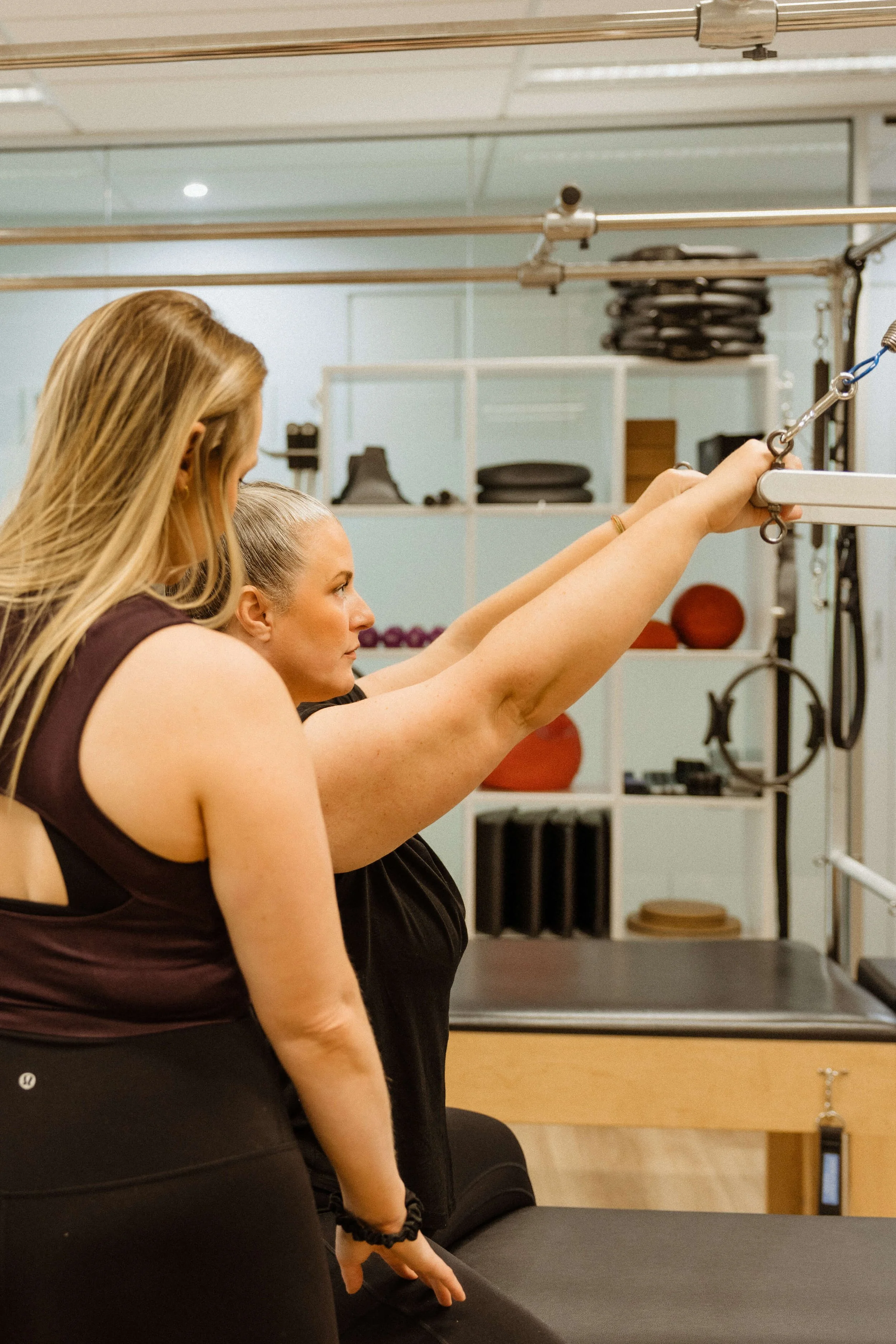Two women in a fitness studio, one assisting the other with a pulley exercise, with gym equipment visible in the background.