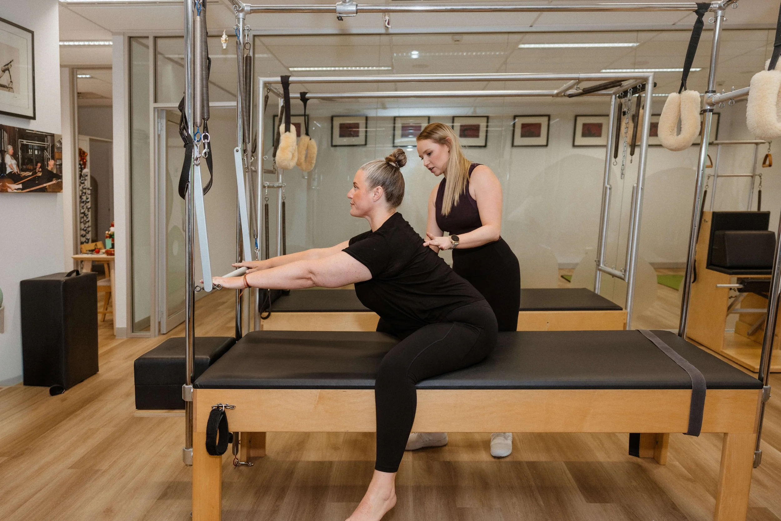 A woman is doing physical therapy on a reformer Pilates machine while a therapist assists her in a fitness studio.
