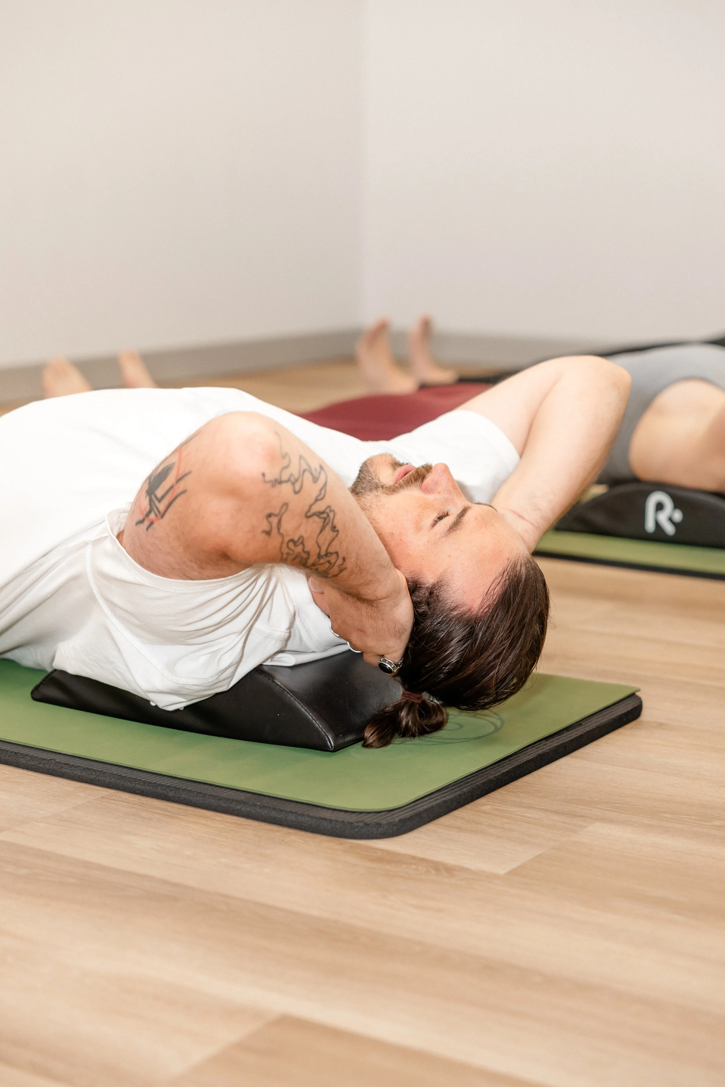 A man with tattoos lying on a yoga mat with his arms behind his head, appearing to be in a relaxed or stretching position in a room.
