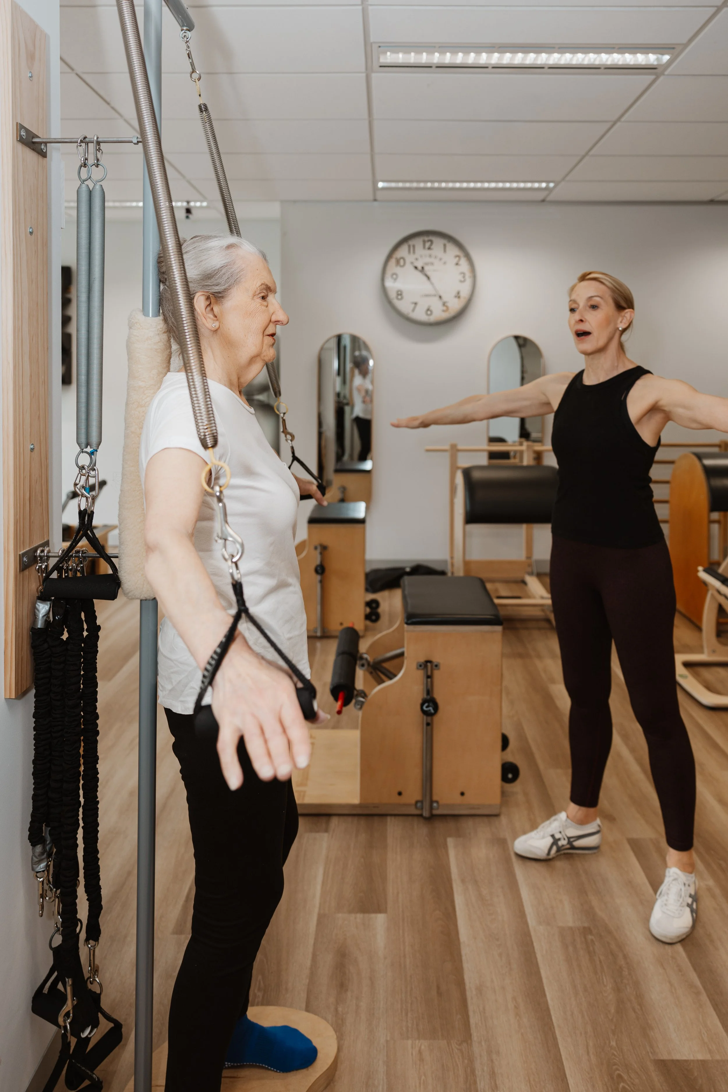 A physical therapy session with an elderly woman standing on a balance platform while a female therapist guides her in an exercise. The woman uses resistance bands attached to a wall, and the therapist extends her arms as she instructs. The room has 
