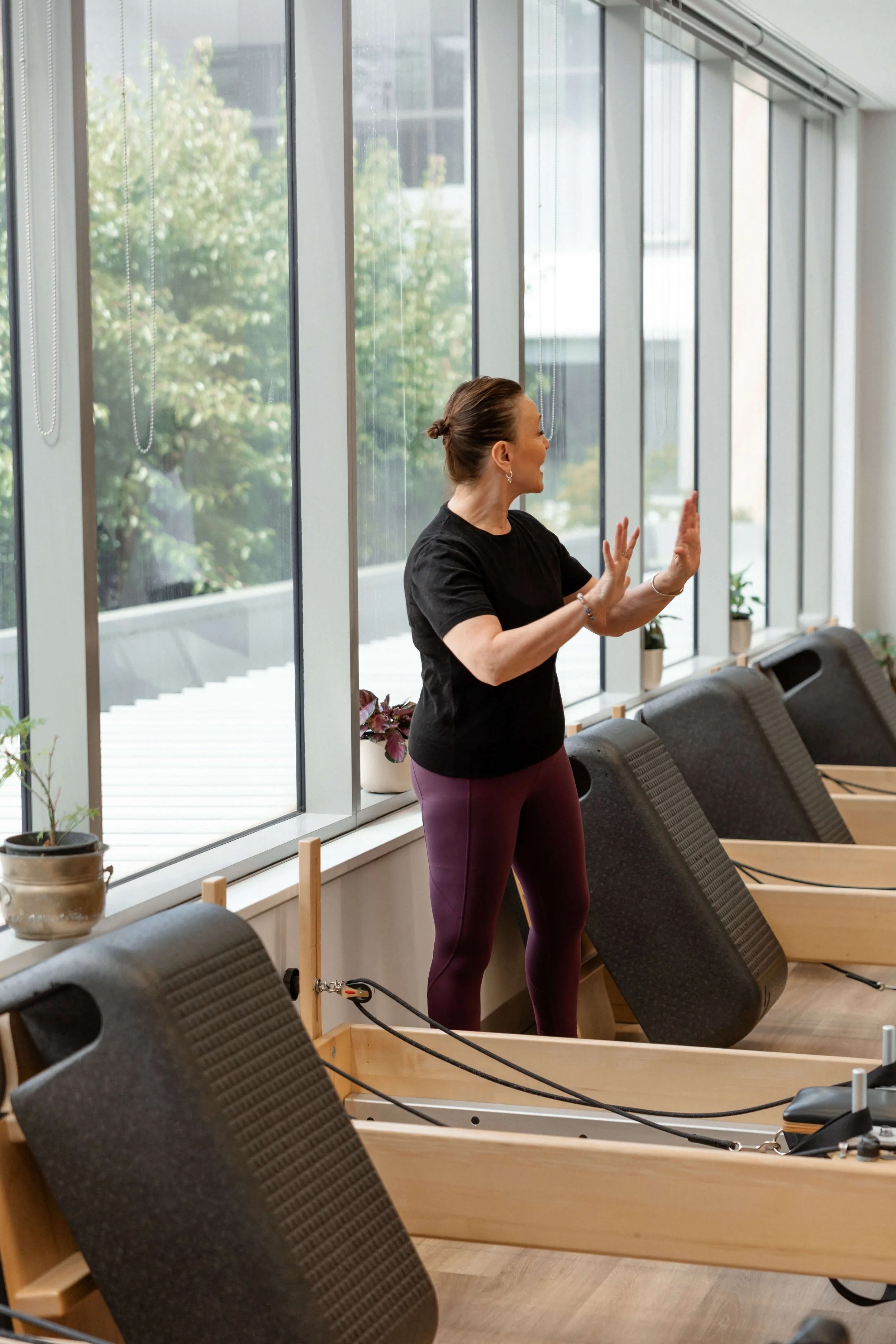 Woman practicing Pilates on reformer machine in bright studio with large windows and potted plants.