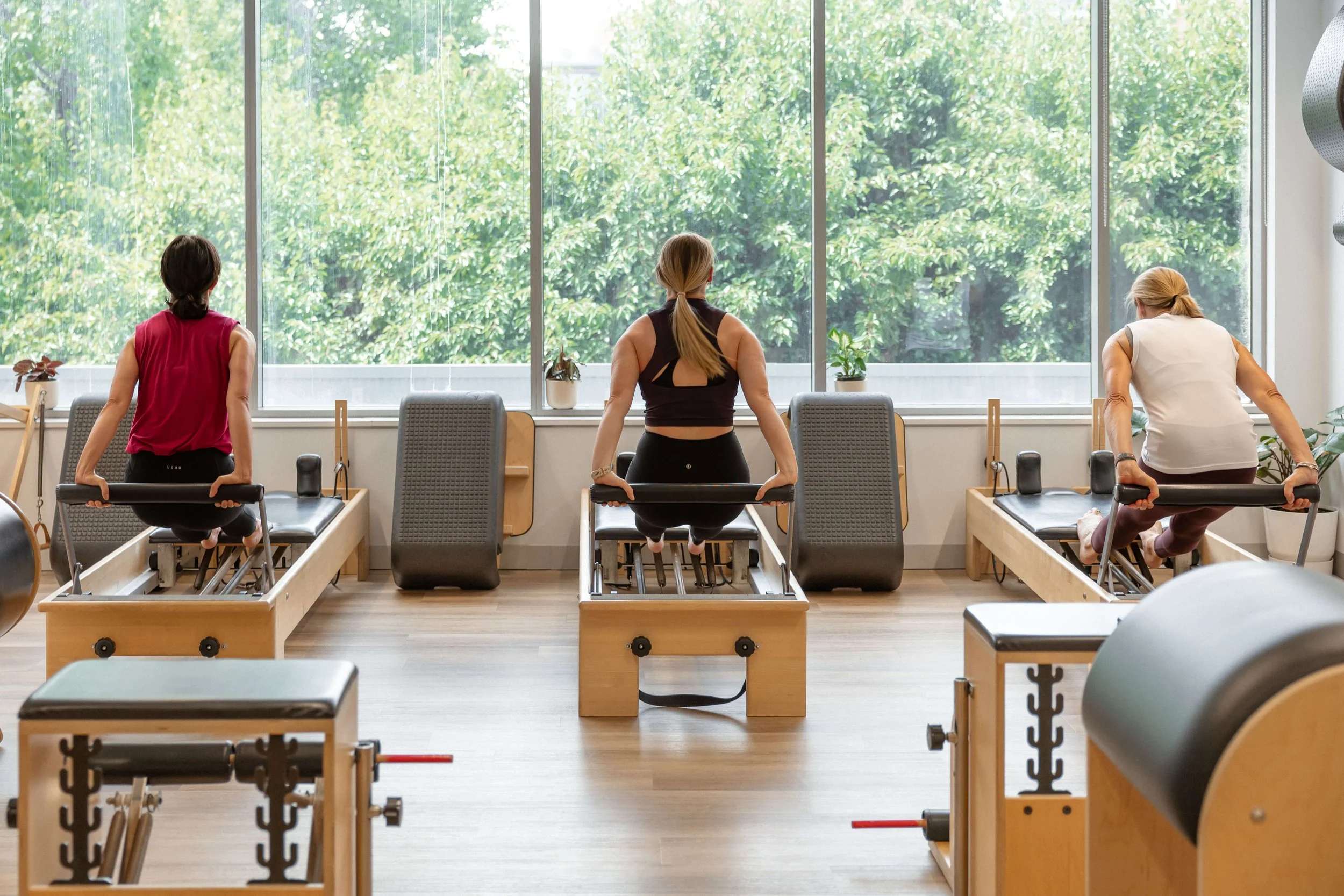 Three women in workout attire using reformer machines in a well-lit fitness studio with large windows and green foliage outside.