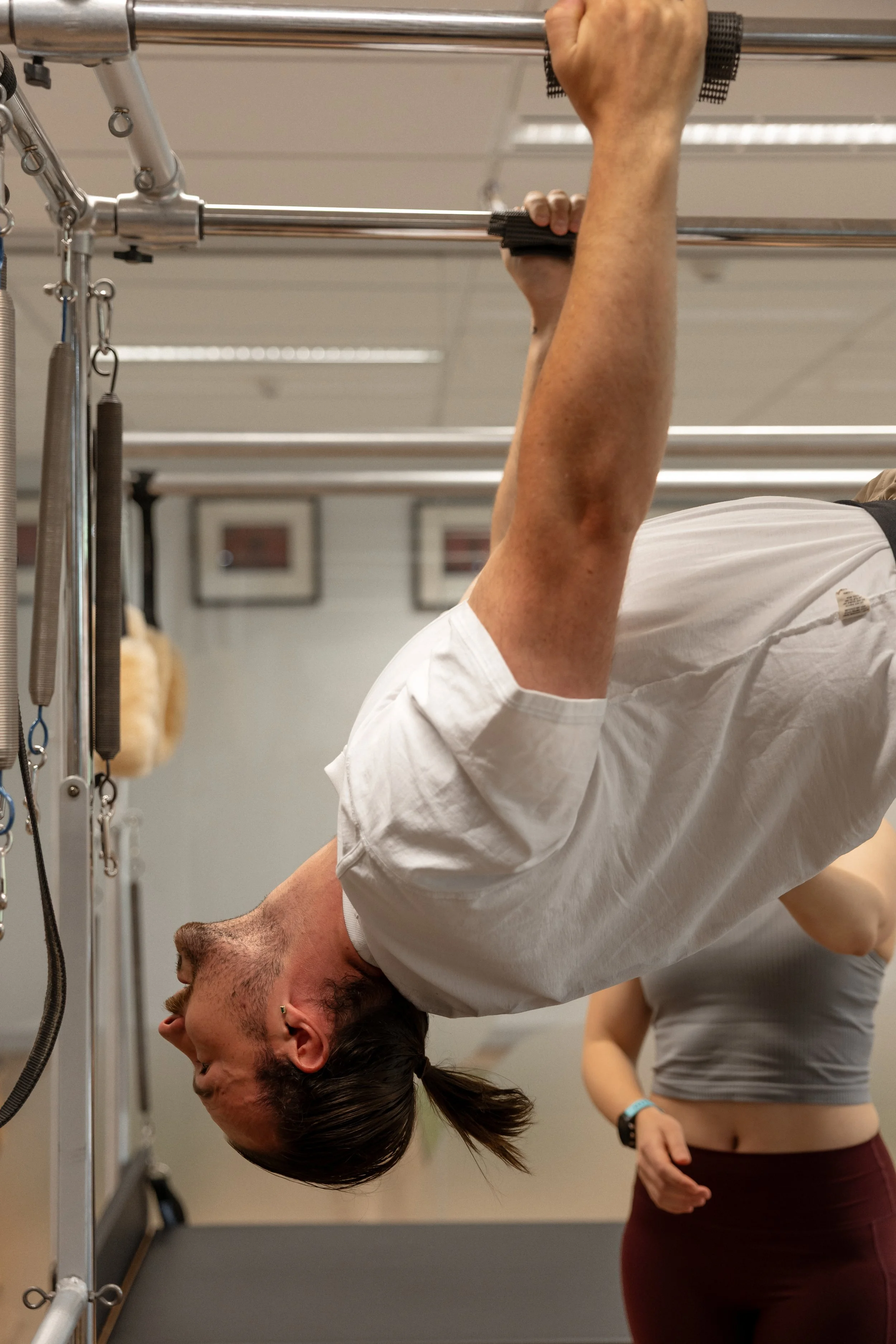 A man is hanging upside down from a pull-up bar, holding onto it with both hands, in a gym. A woman in workout clothes is standing nearby with her hands on her hips.