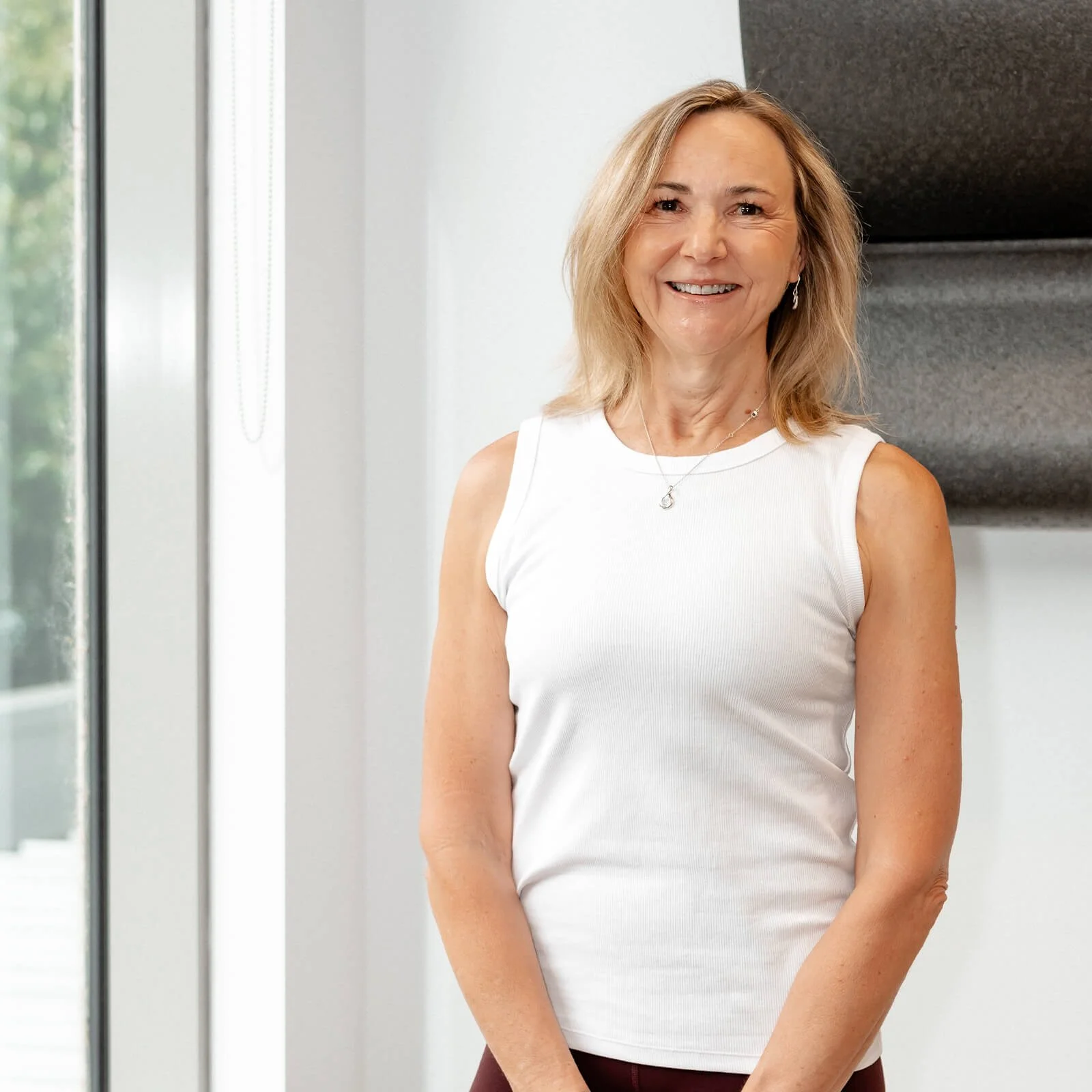 A middle-aged woman with blonde hair and a white sleeveless top smiling indoors near a large window and a black textured wall.