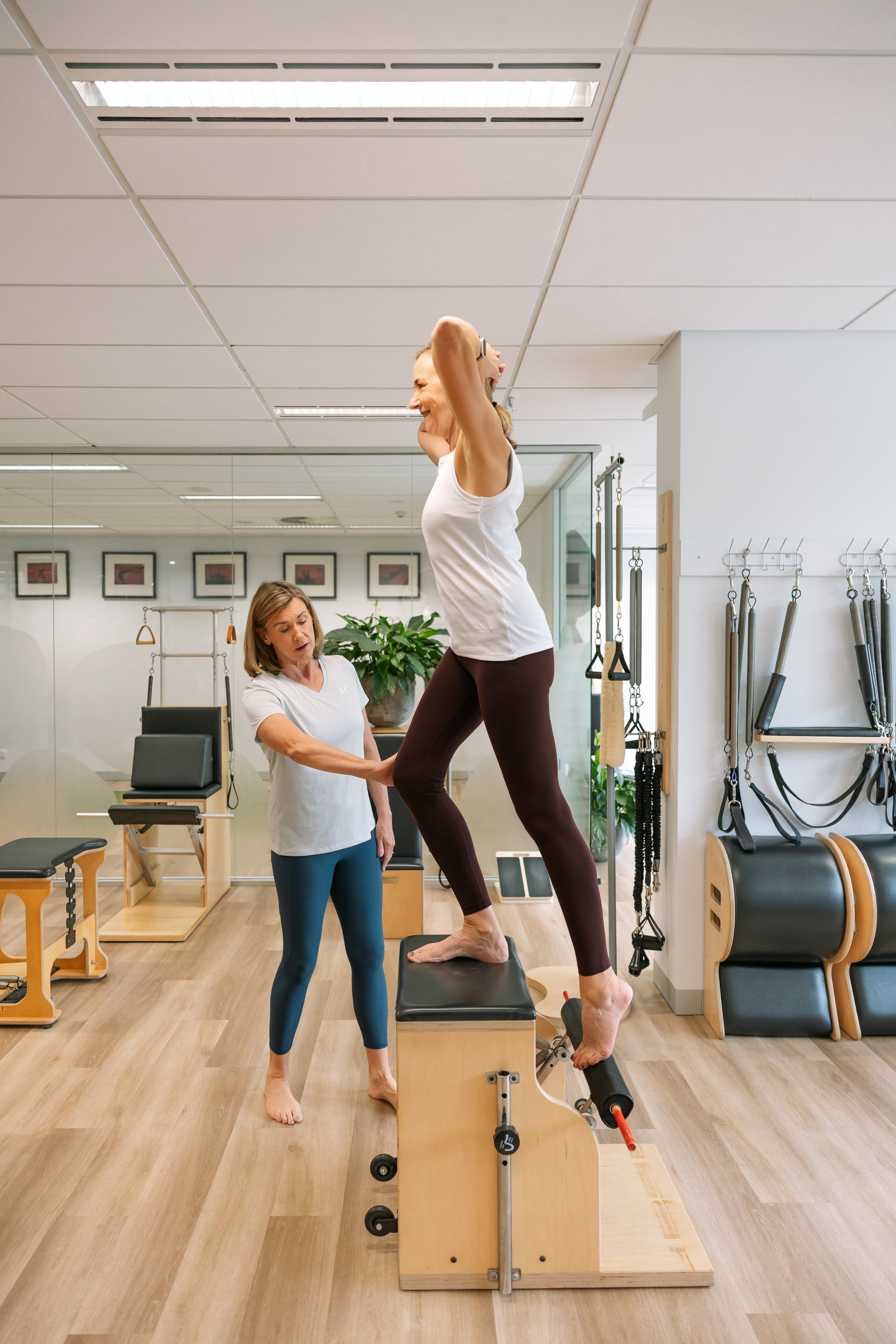 A woman performs a Pilates exercise on a specialized reformer machine in a Pilates studio, assisted by a trainer.