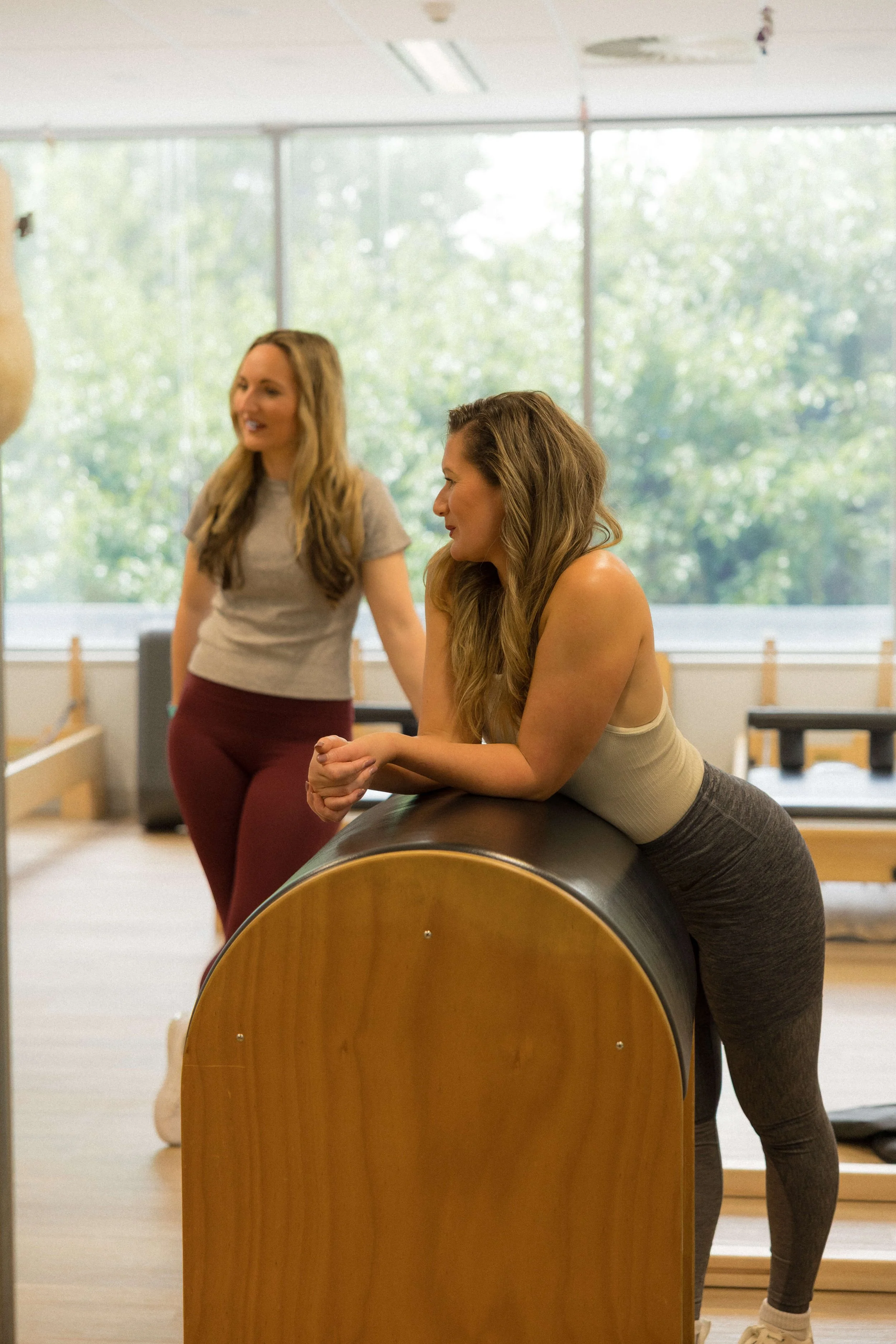 Two women in a fitness or physical therapy setting, one leaning on a padded exercise barrel and the other standing behind her, engaging in conversation.