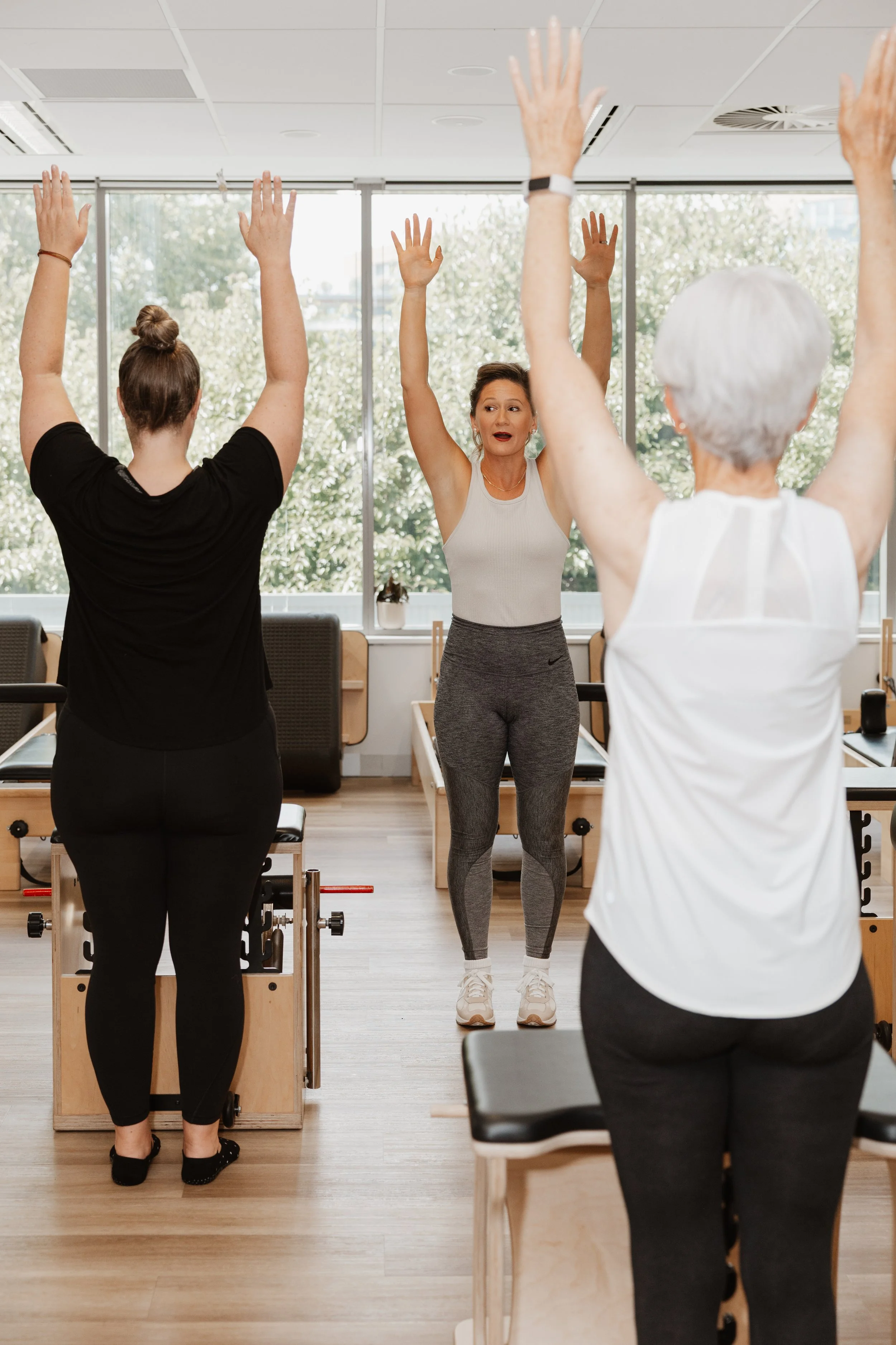 Three women participating in a seated exercise class, with their arms raised overhead, in a bright room with large windows and natural light.