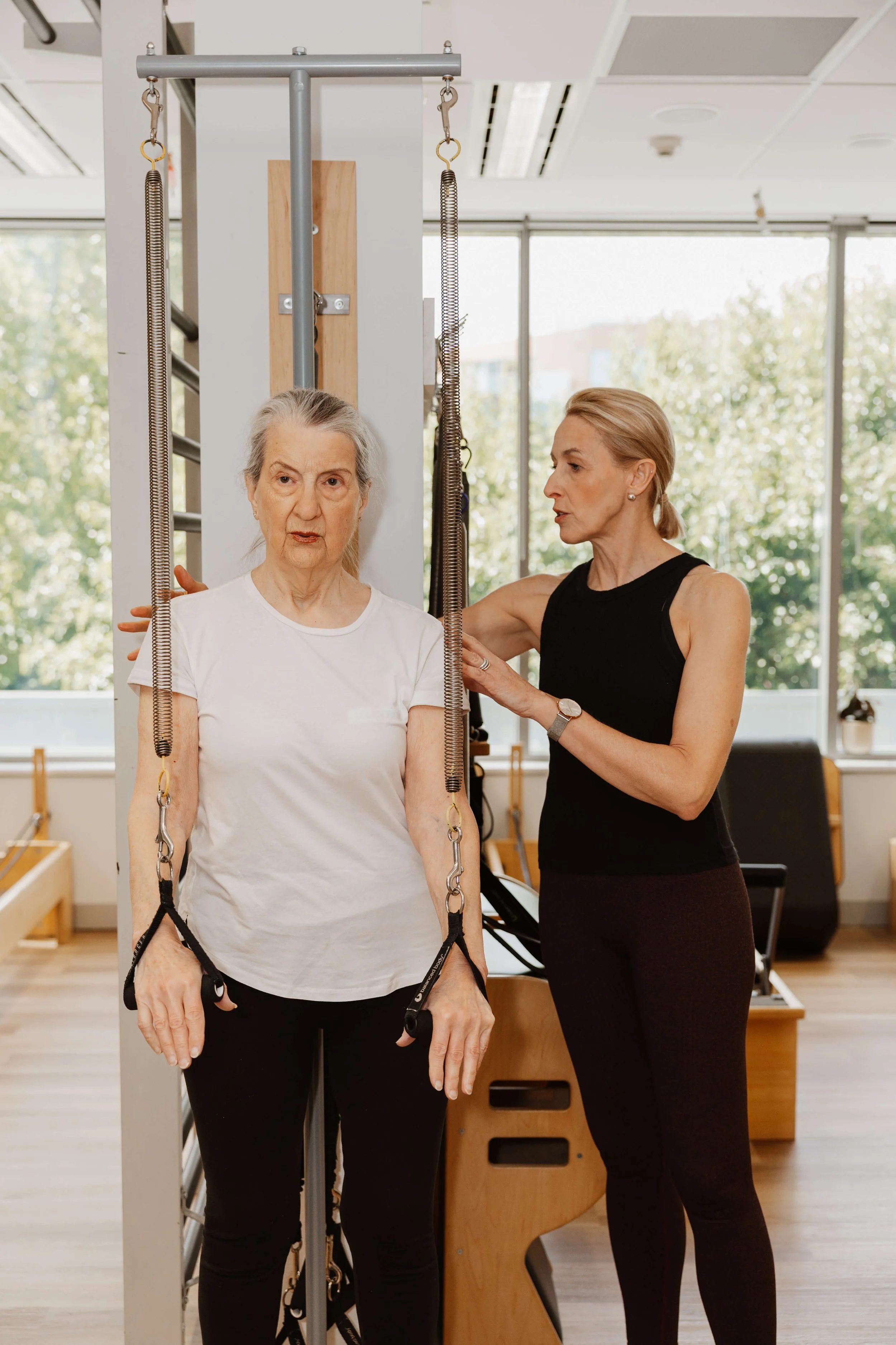 A physical therapist assists an elderly woman during a rehabilitation session using a suspension trainer in a well-lit clinic with large windows.