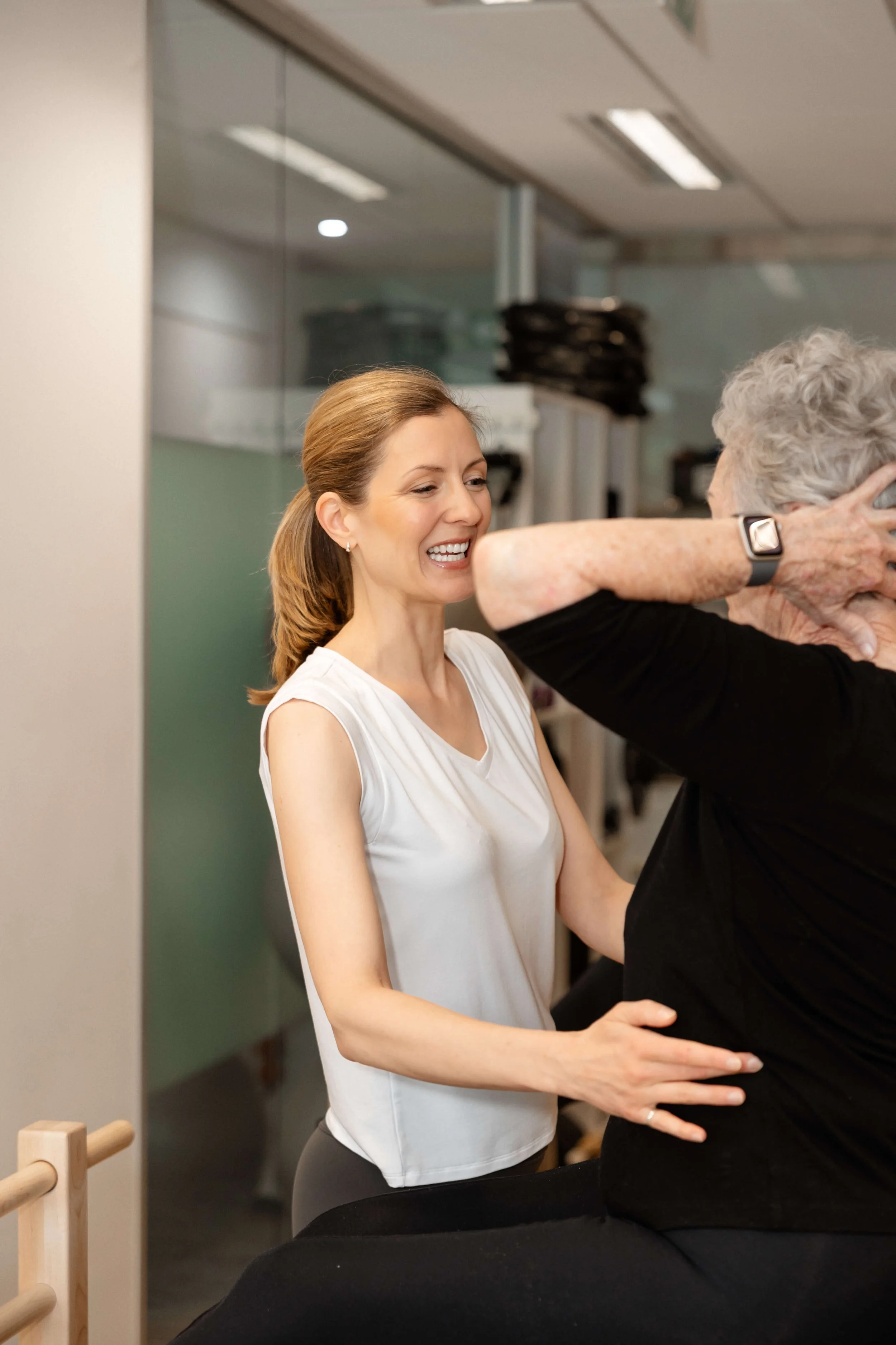 Fitness instructor assisting an elderly woman with stretching exercises in a gym or fitness studio.