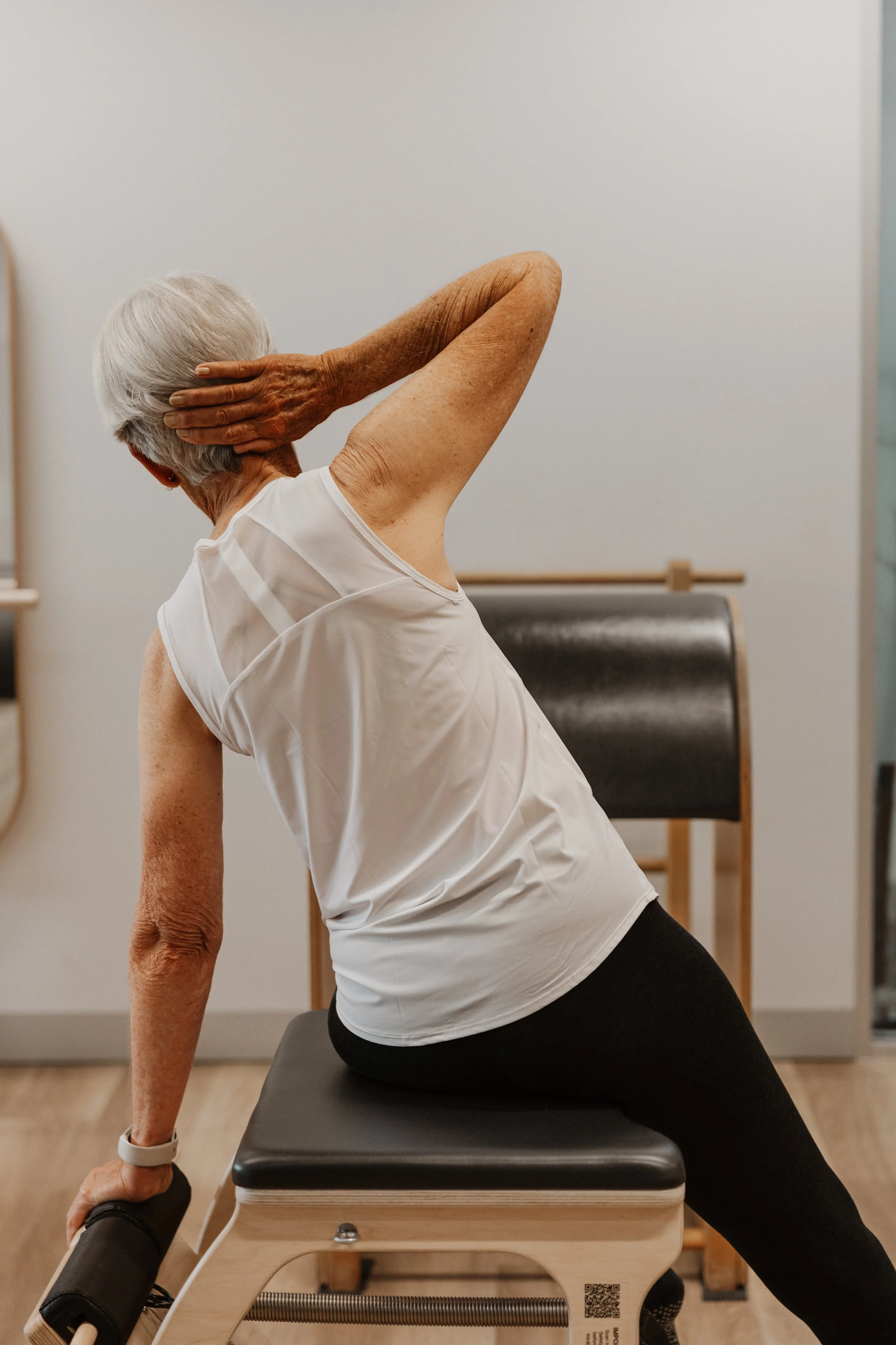 An elderly woman with gray hair performing a stretching exercise while seated on a Pilates reformer machine, with her left hand behind her head and her right arm supporting her on the machine.