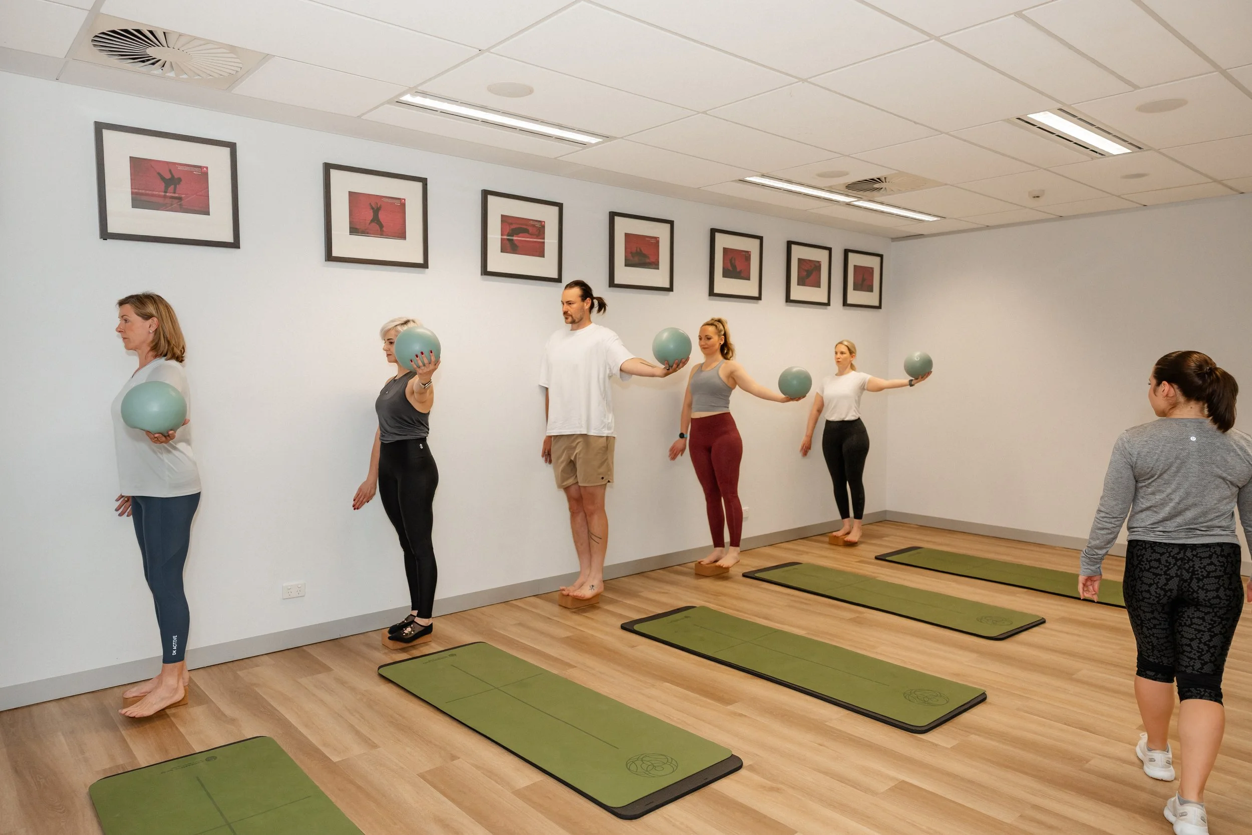 People in a fitness class holding exercise balls and balancing on mats in a studio.