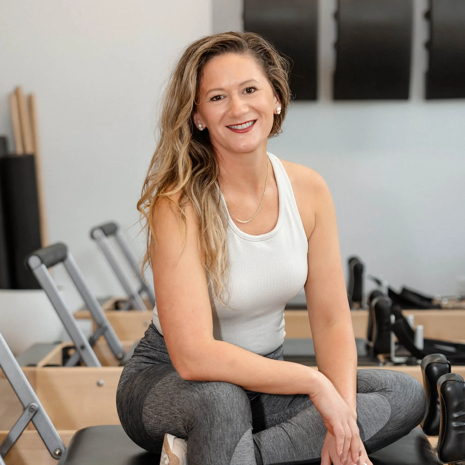 A woman with long, wavy blonde hair smiling, sitting on a fitness step in a gym, wearing a white tank top and gray leggings.