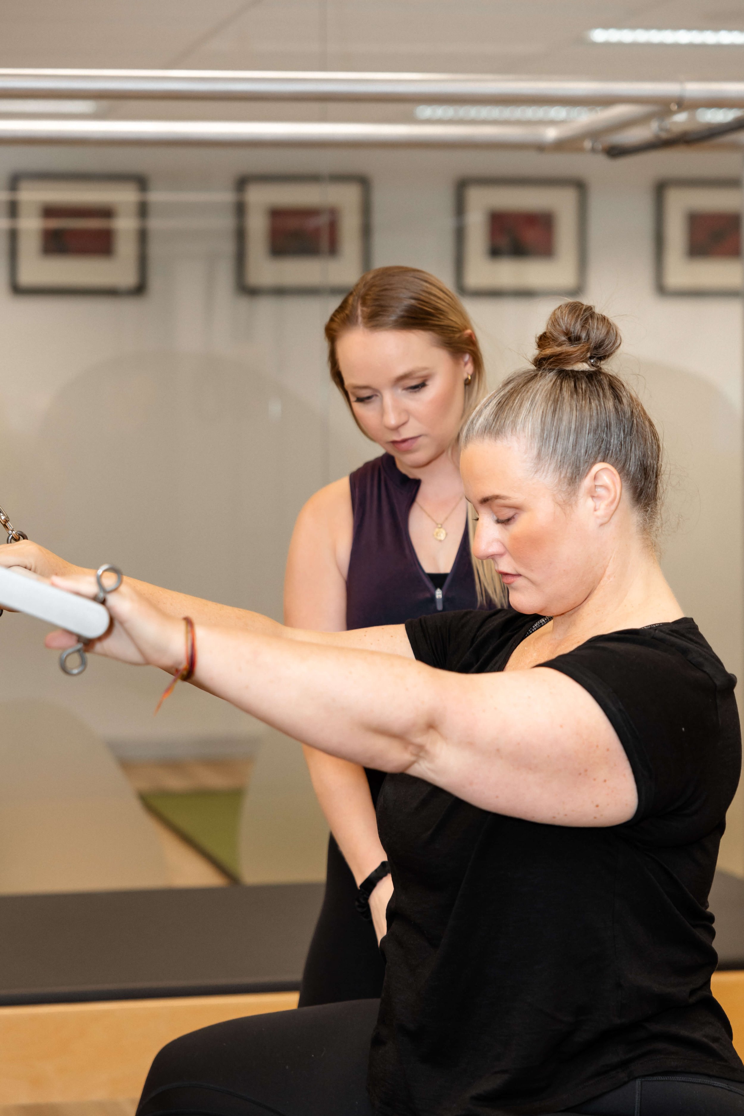 A woman in a black shirt performs an exercise with resistance bands while another woman in a sleeveless top assists and watches in a fitness or physical therapy setting.