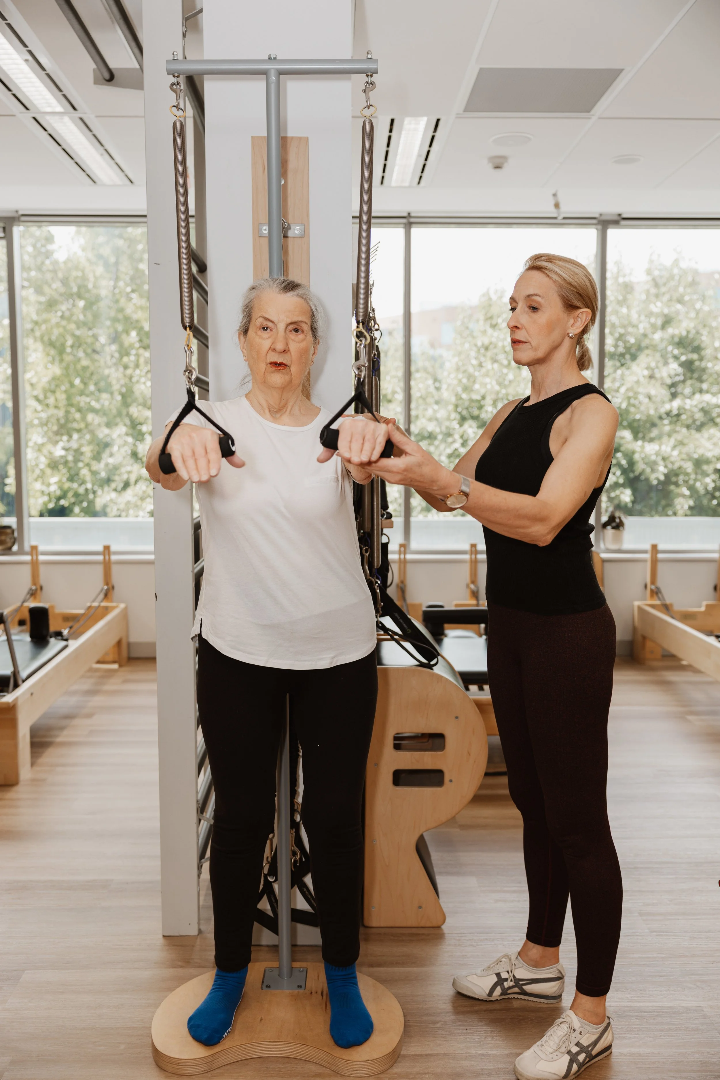 Two women exercising with suspension straps in a gym, one elderly woman and one middle-aged woman assisting her.