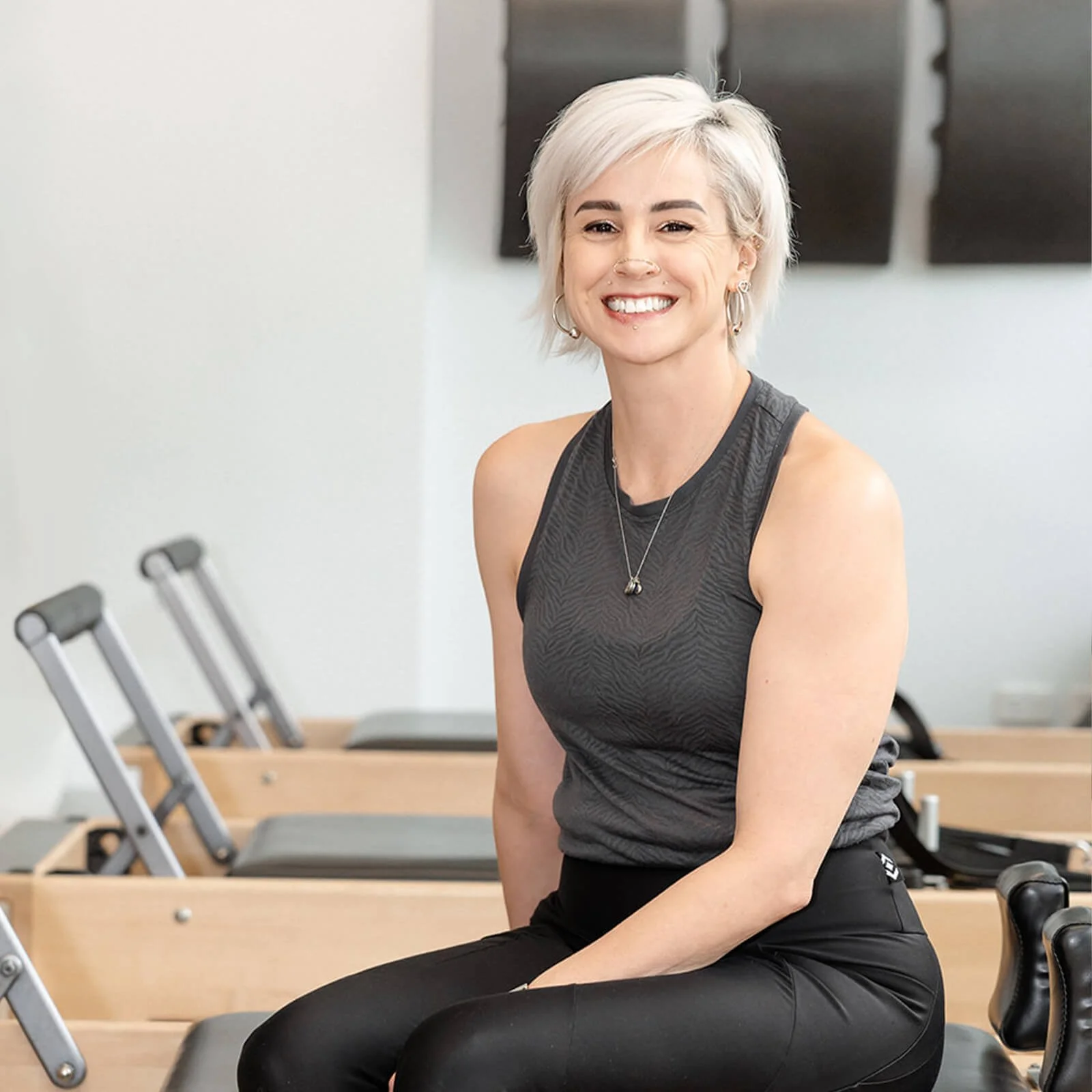 A smiling woman with short, platinum blonde hair sitting in a fitness or Pilates studio.