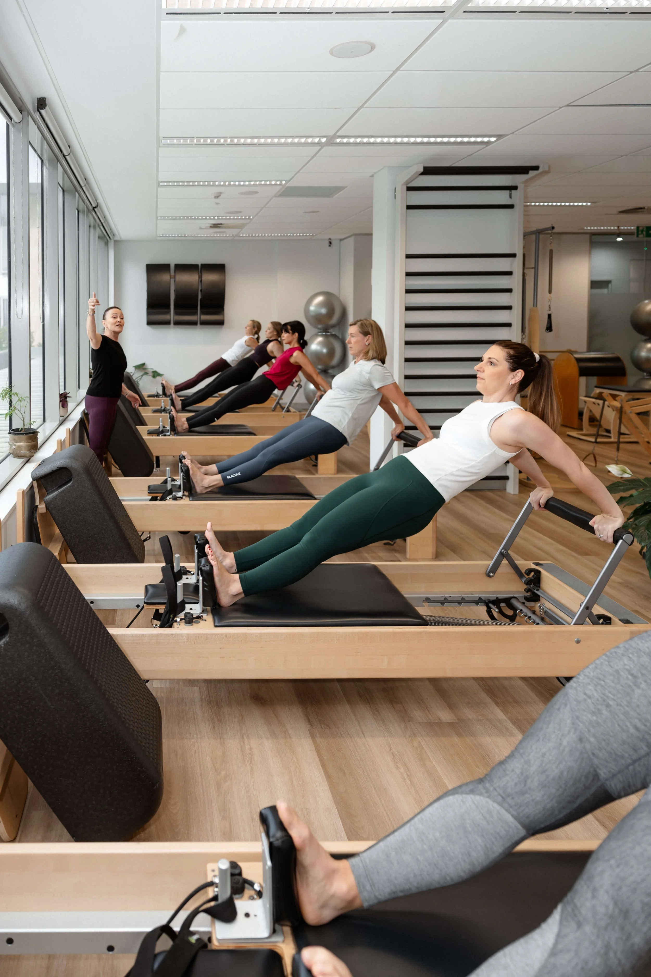 A fitness instructor leads a group class in a gym with several women using Pilates reformer machines for exercise.