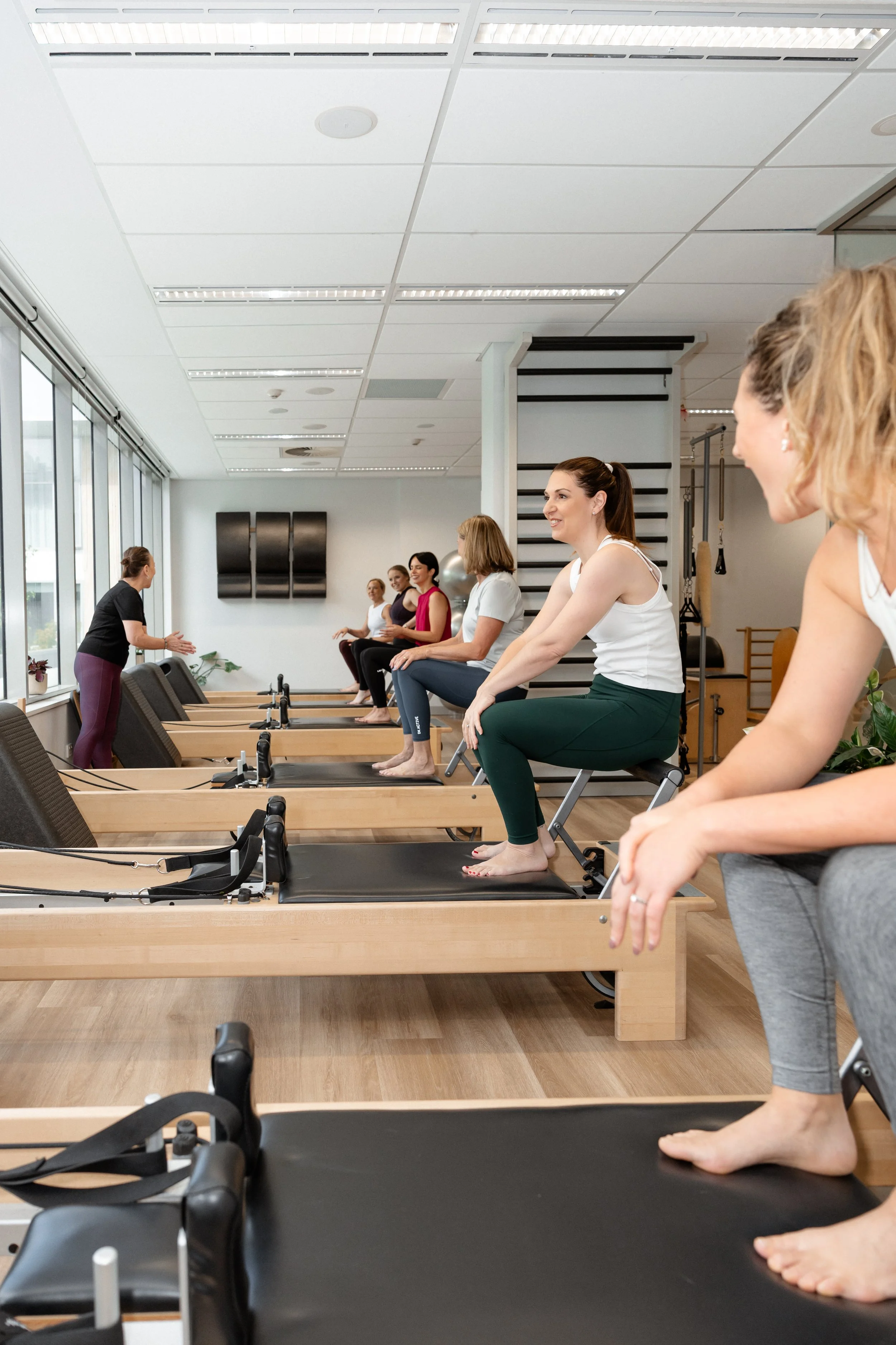 A group fitness class in a gym with women sitting on reformer machines, instructor demonstrating at the front.